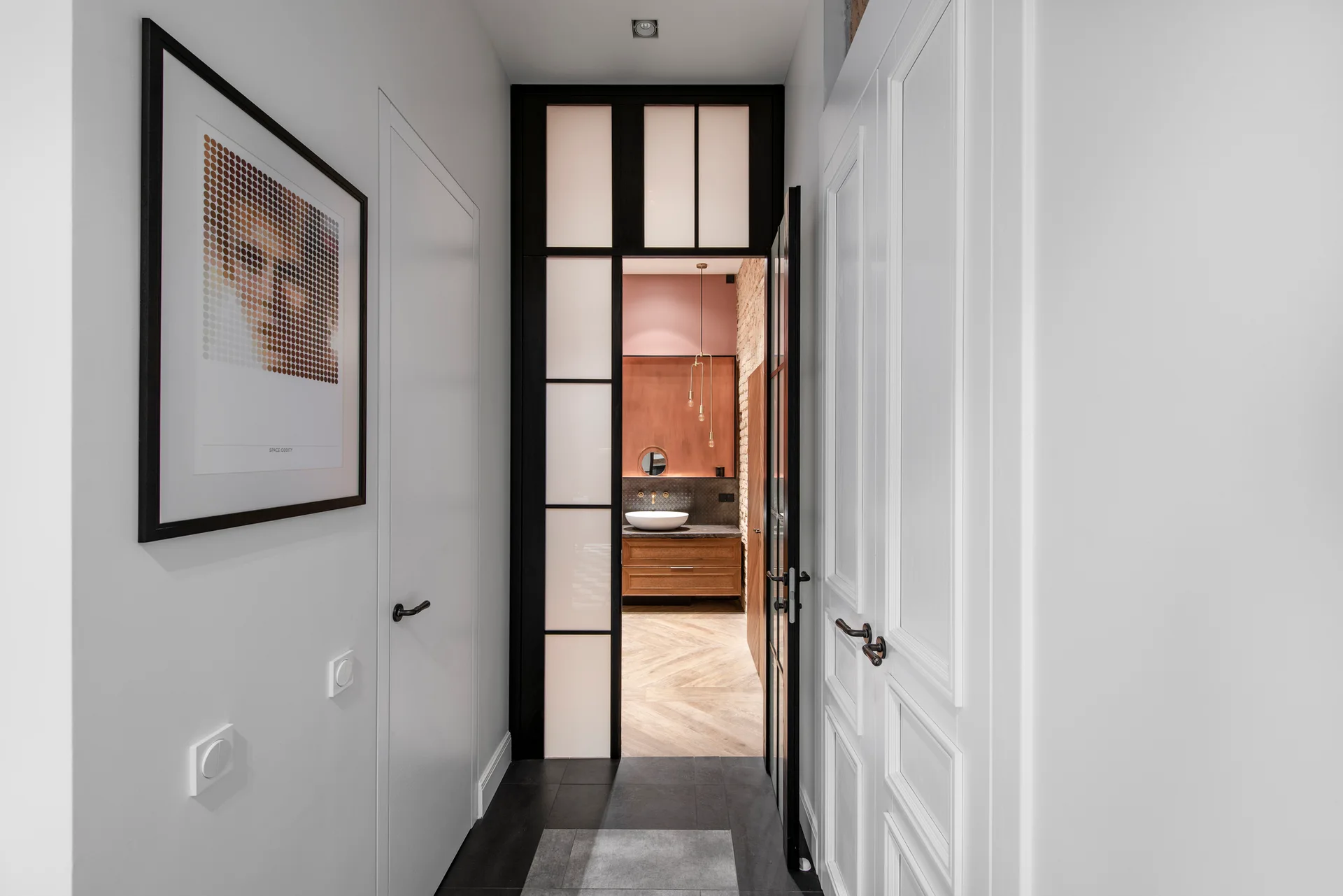 A white hallway with dark tile floors looks through a black-framed glass door into a bathroom featuring a wooden vanity and exposed brick wall.