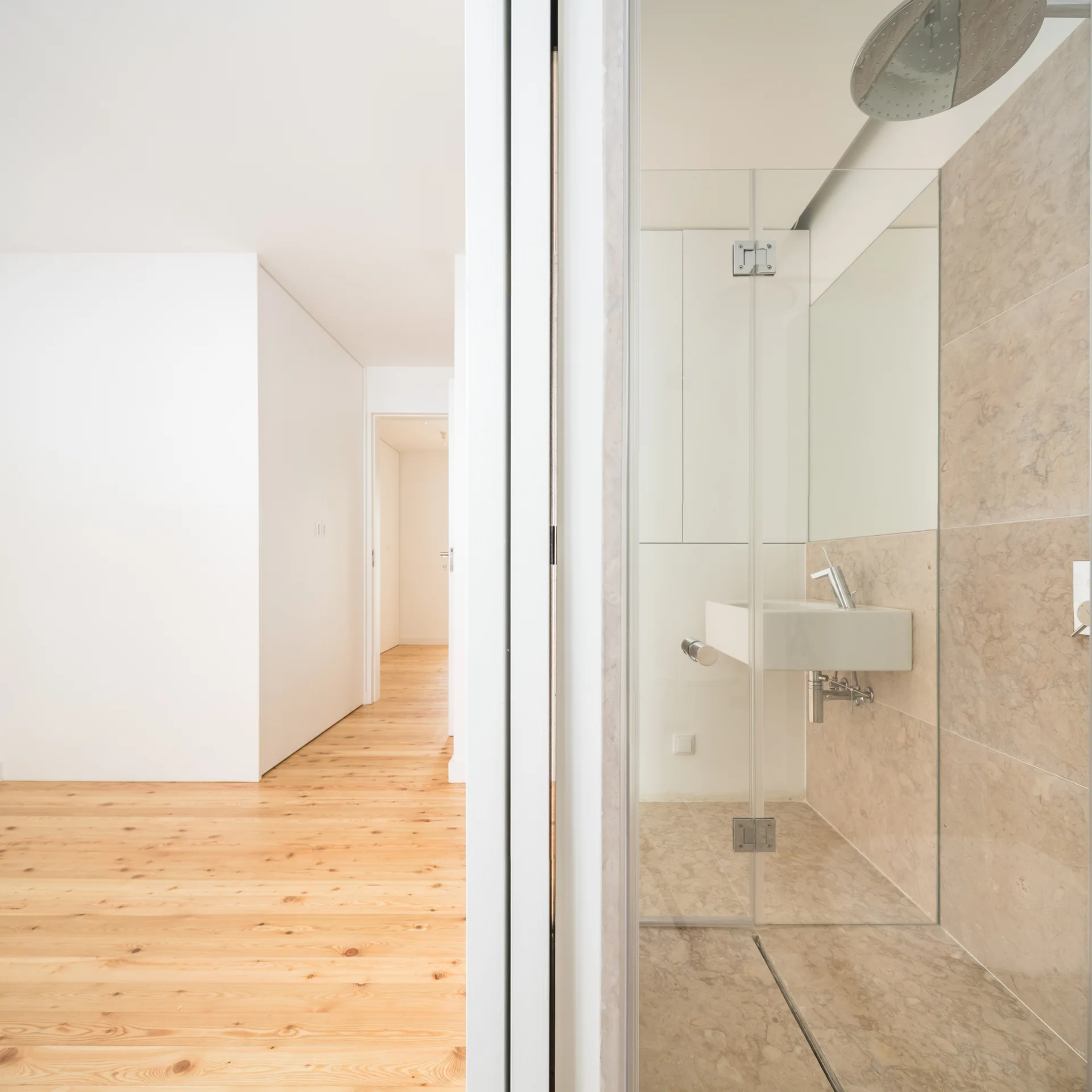 A minimalist bathroom with beige stone tiles and a glass shower next to a white-walled hallway with light wood floors.