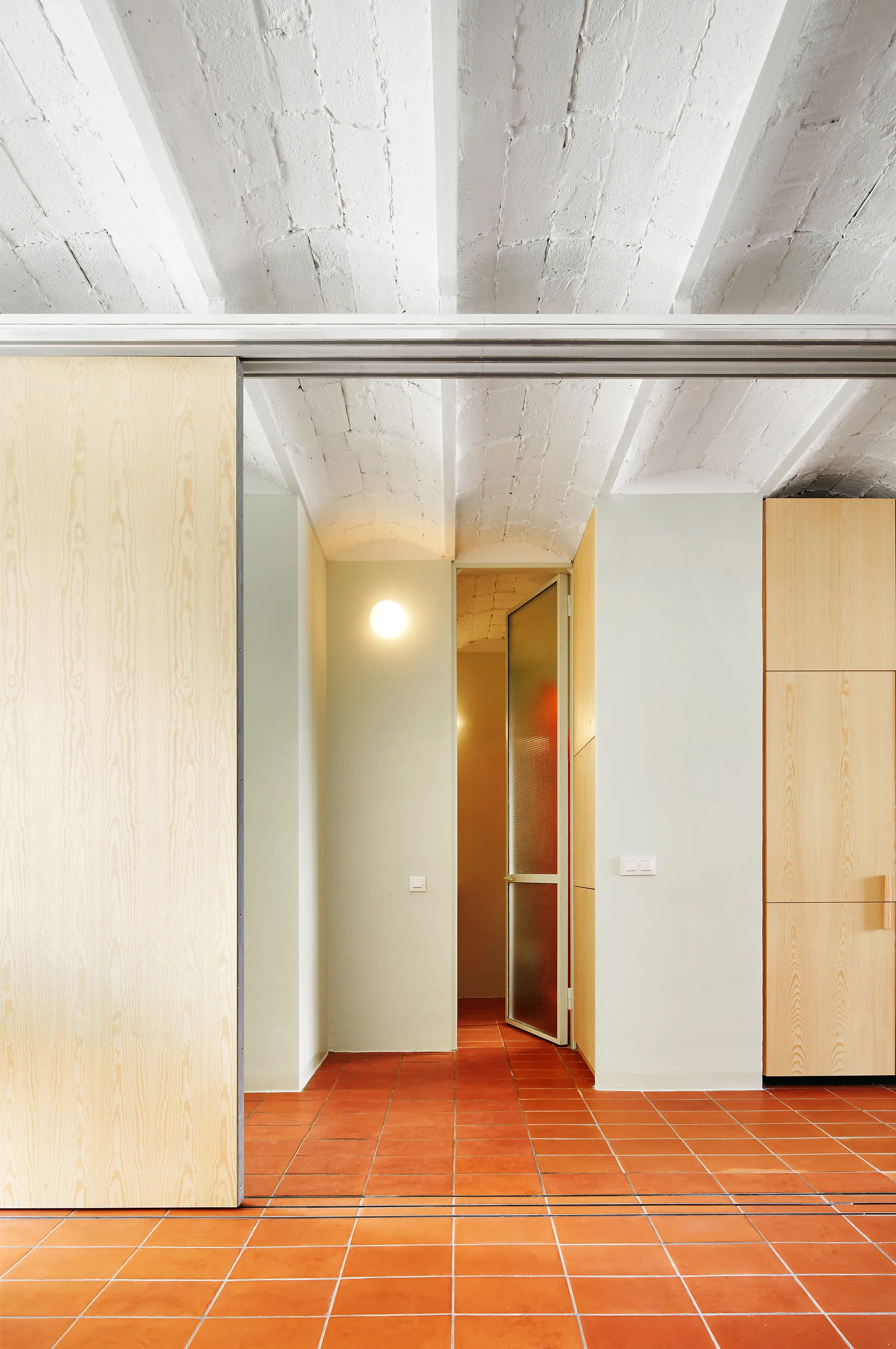 A modern hallway features a white painted vaulted brick ceiling, red terra cotta floor tiles, and a sliding light wood panel door.