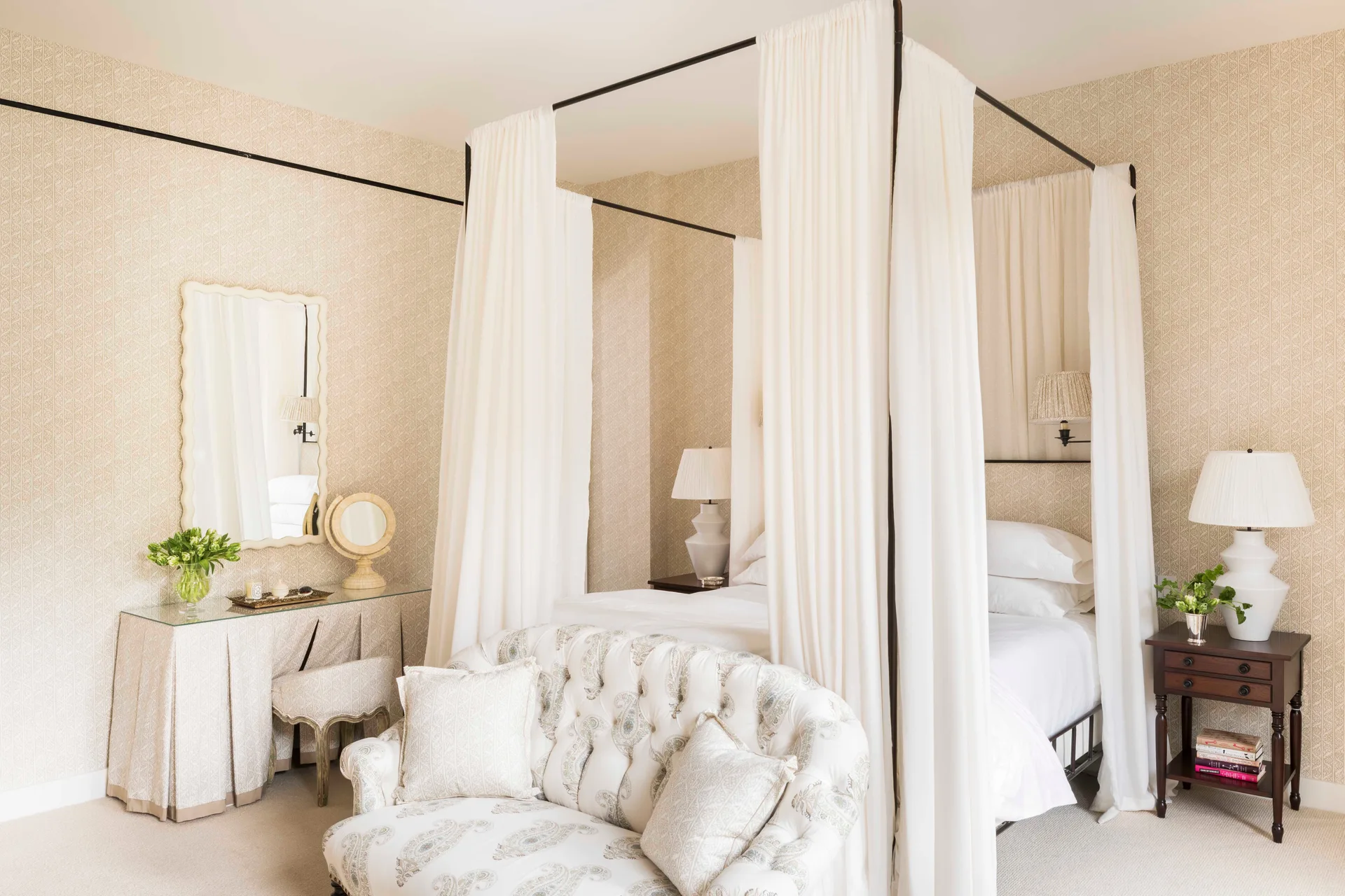 A beige bedroom featuring a four-poster canopy bed with white drapes, a tufted patterned sofa, and a skirted vanity table with a scalloped mirror.