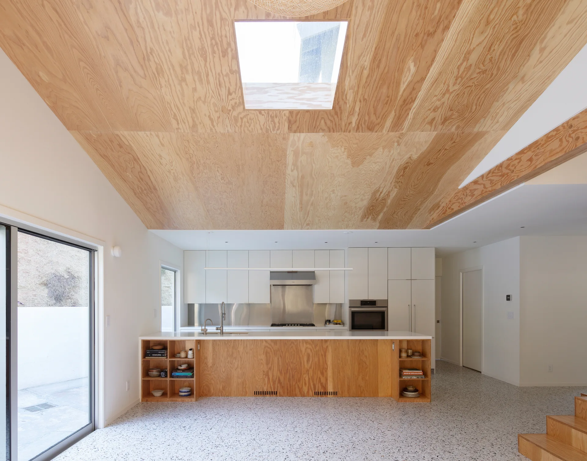 A modern kitchen features a vaulted plywood ceiling with a skylight, a wood-clad island, and speckled terrazzo flooring.