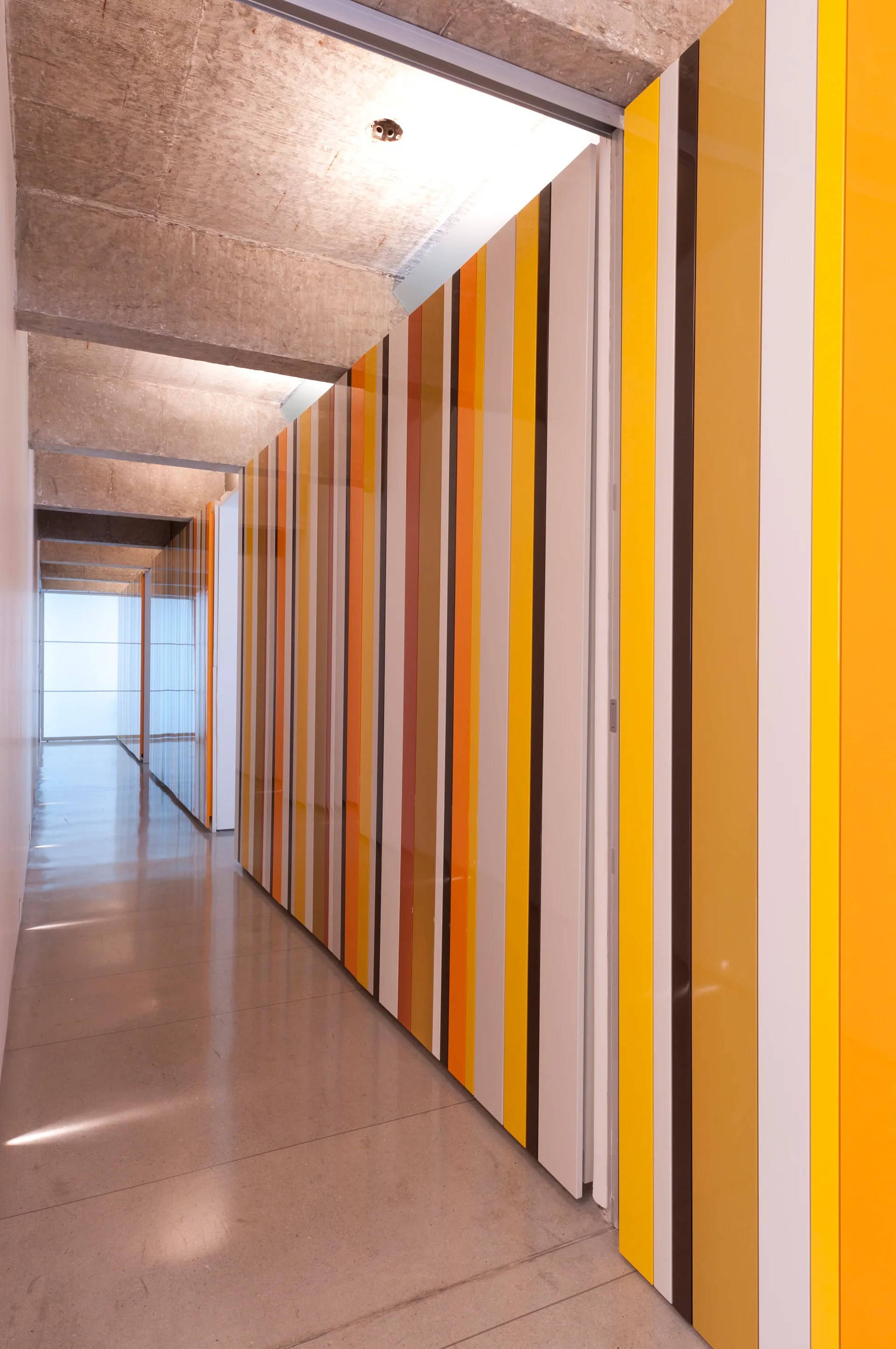 A long hallway features raw concrete ceiling beams, polished grey floors, and a wall of glossy yellow, orange, and brown vertical striped panels.