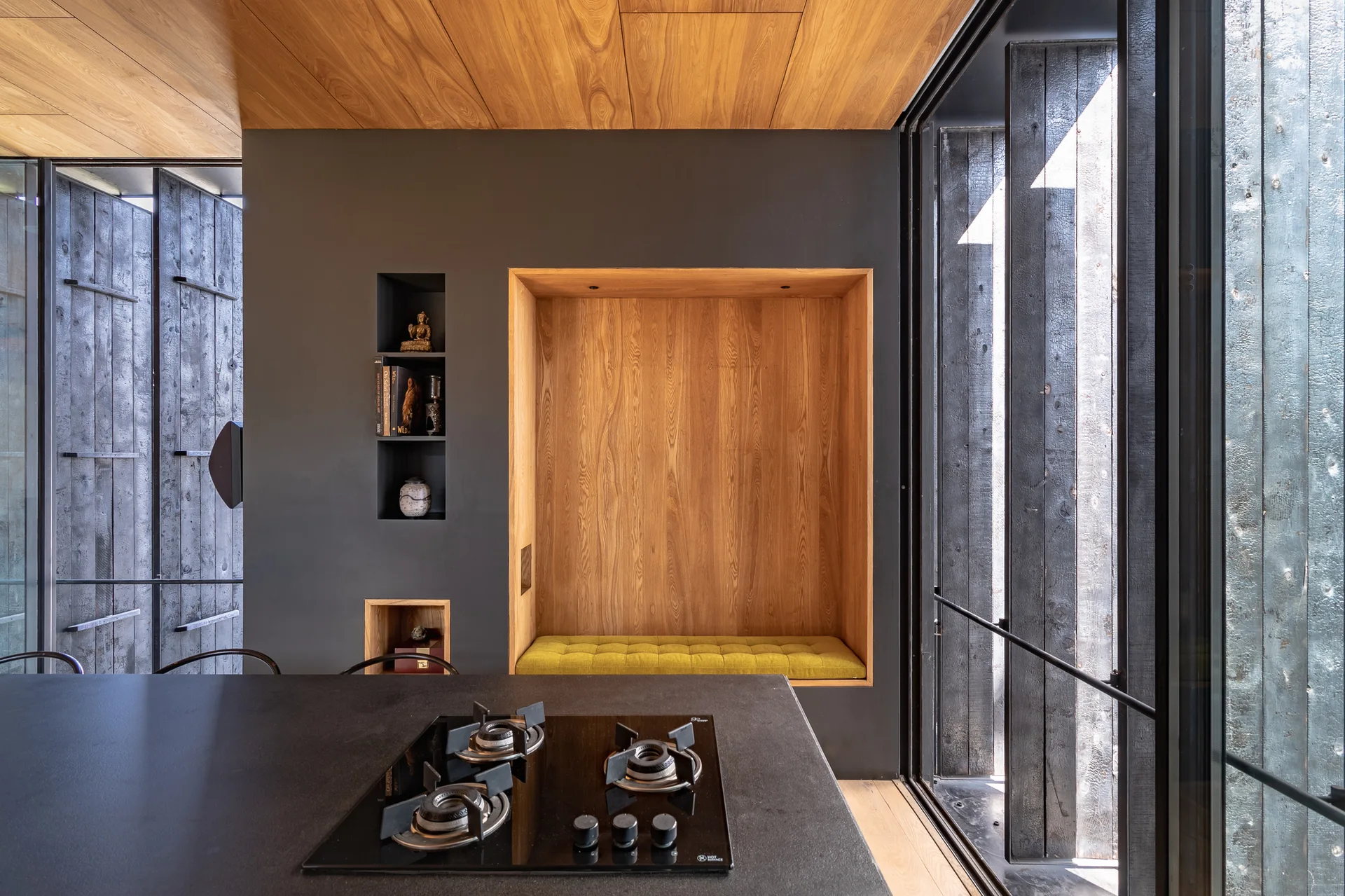 A modern kitchen featuring a black stovetop island in front of a charcoal gray wall with a recessed wood-paneled bench and mustard yellow cushion.