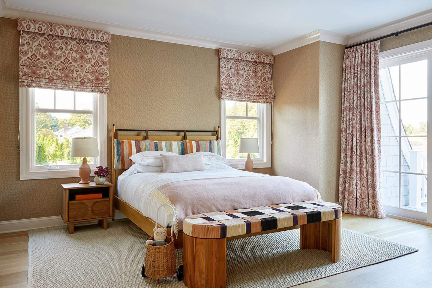A transitional bedroom featuring a wooden bed with a striped headboard, a woven bench, pink patterned roman shades, and two ceramic lamps.