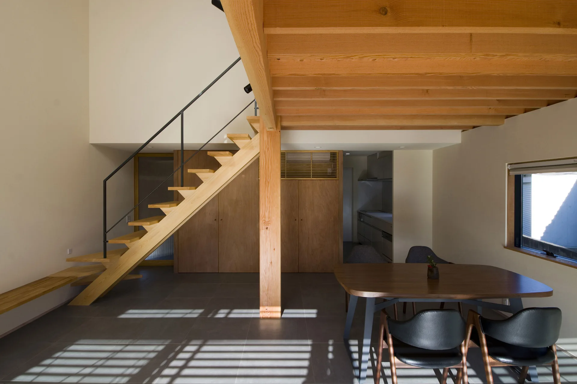 A modern dining room features a dark wood table, black leather chairs, a floating wooden staircase, and exposed ceiling beams on grey tiled floors.