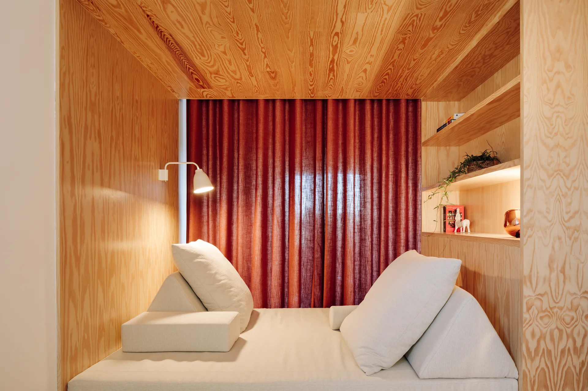 A built-in plywood sleeping nook features an off-white daybed with plush pillows, recessed side shelving, and deep red curtains in the background.