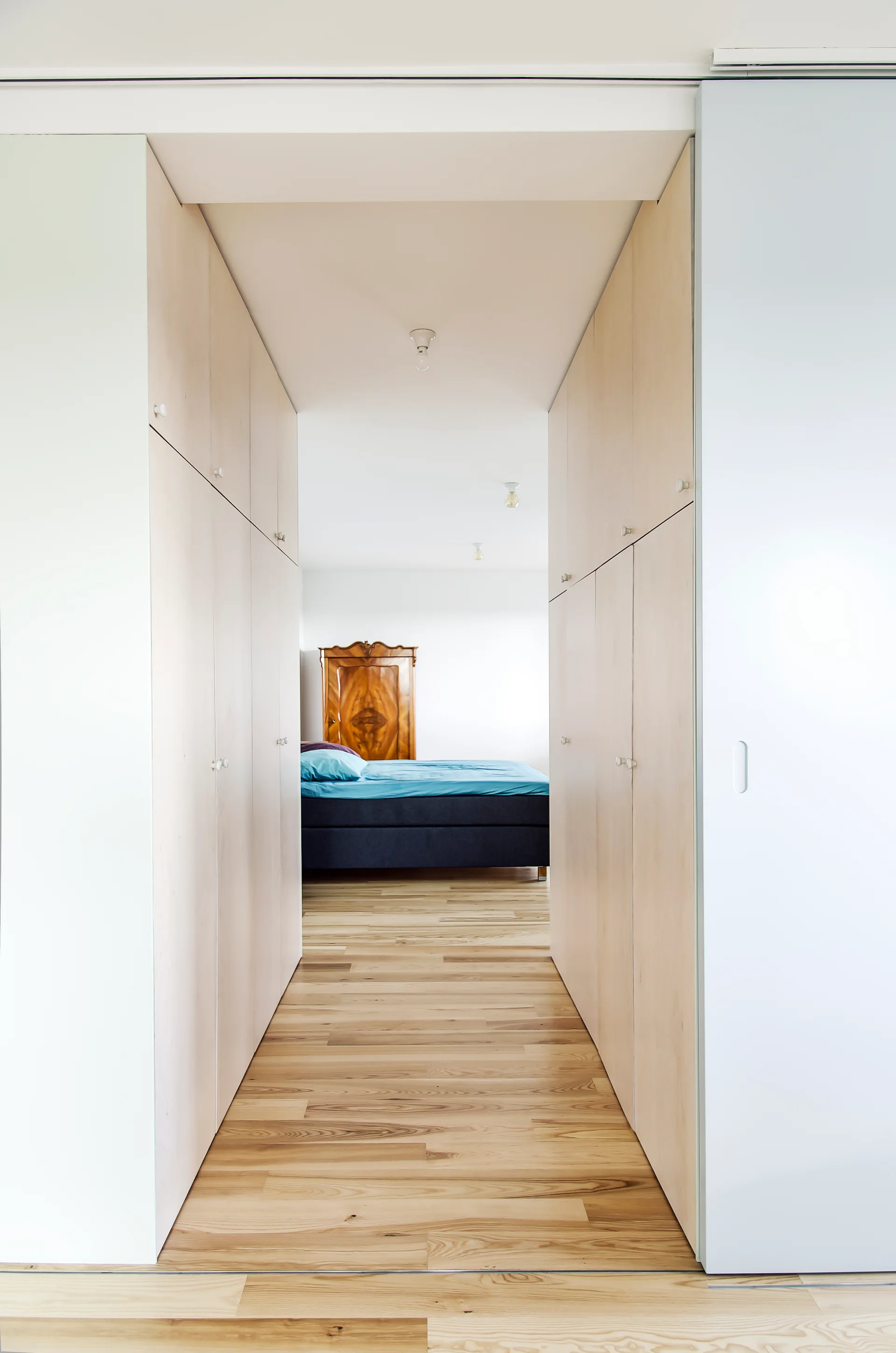 A hallway lined with built-in light wood cabinets and hardwood flooring leads into a white bedroom featuring a blue bed and antique wooden wardrobe.