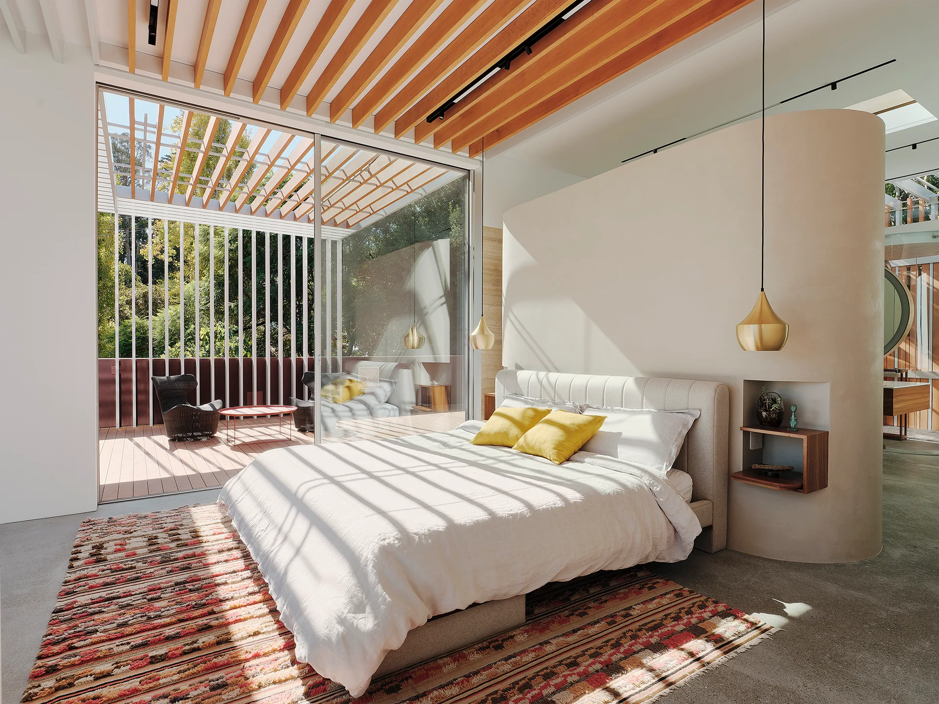 A minimalist bedroom features an upholstered bed, a curved partition wall, and exposed wooden ceiling beams that look out onto a sunny balcony.