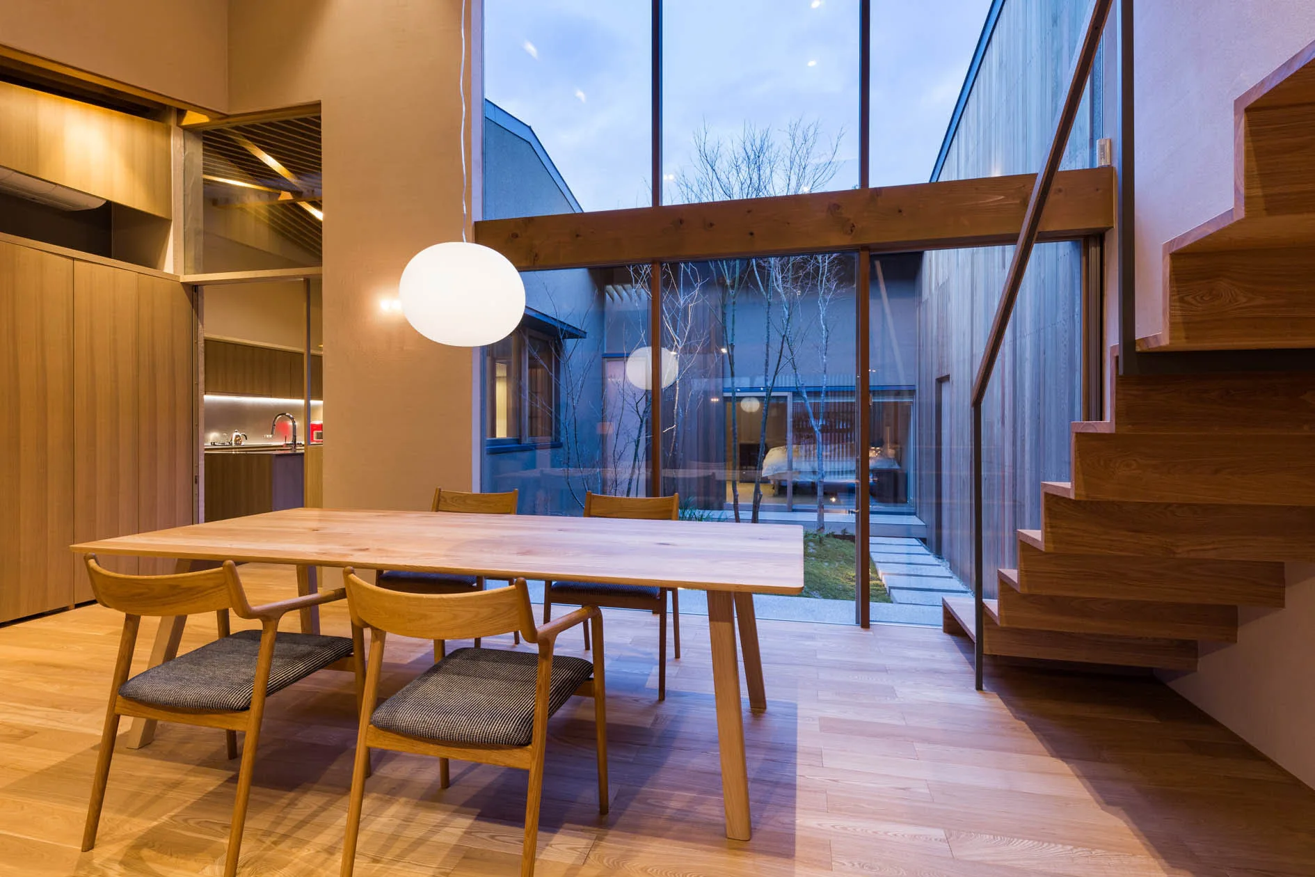 A minimalist dining room featuring a light wood table and chairs next to a floating wooden staircase and floor-to-ceiling windows.