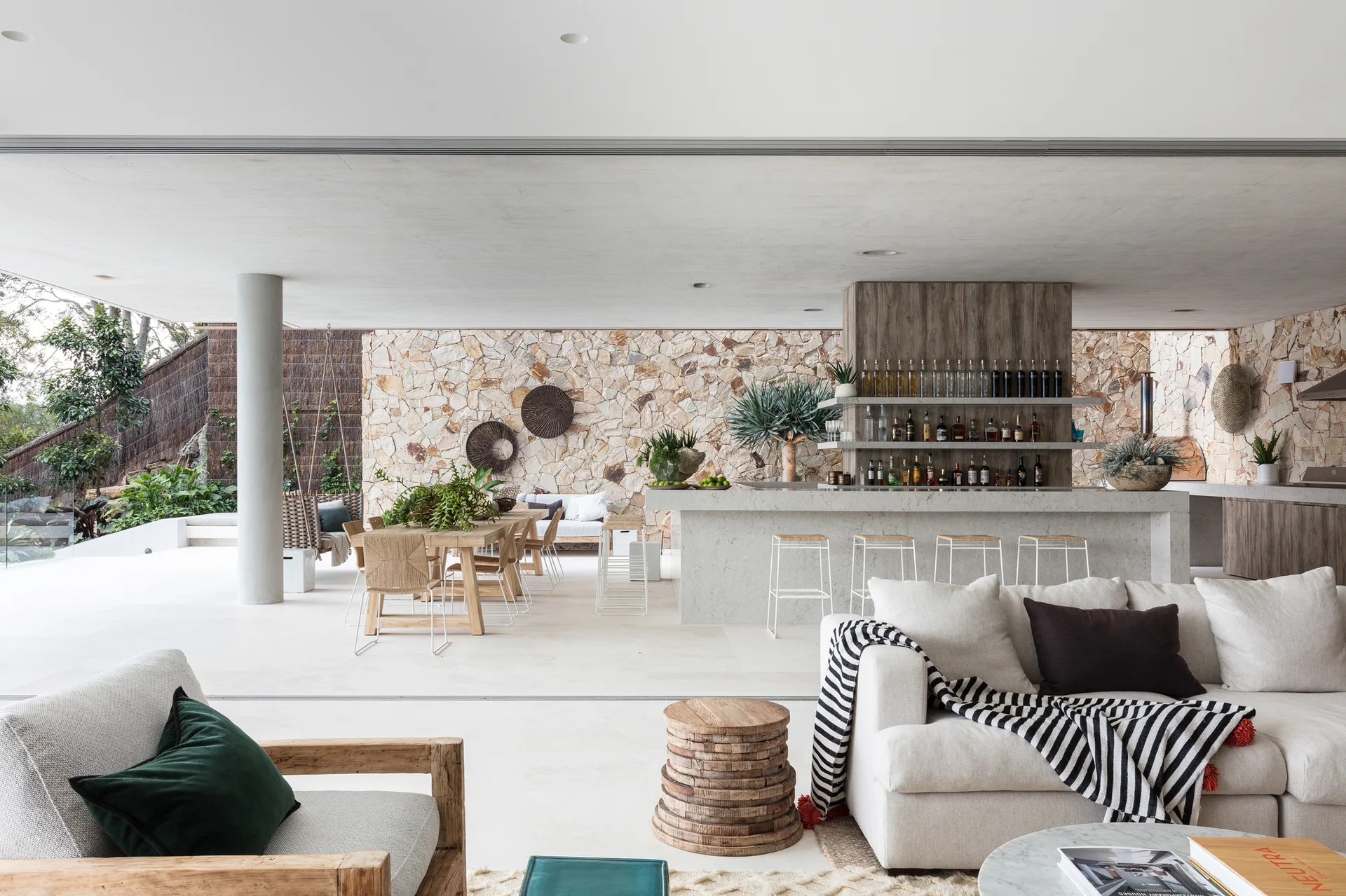 A modern open-air living space featuring a white sofa, a stone bar counter with stools, and a textured stone accent wall.