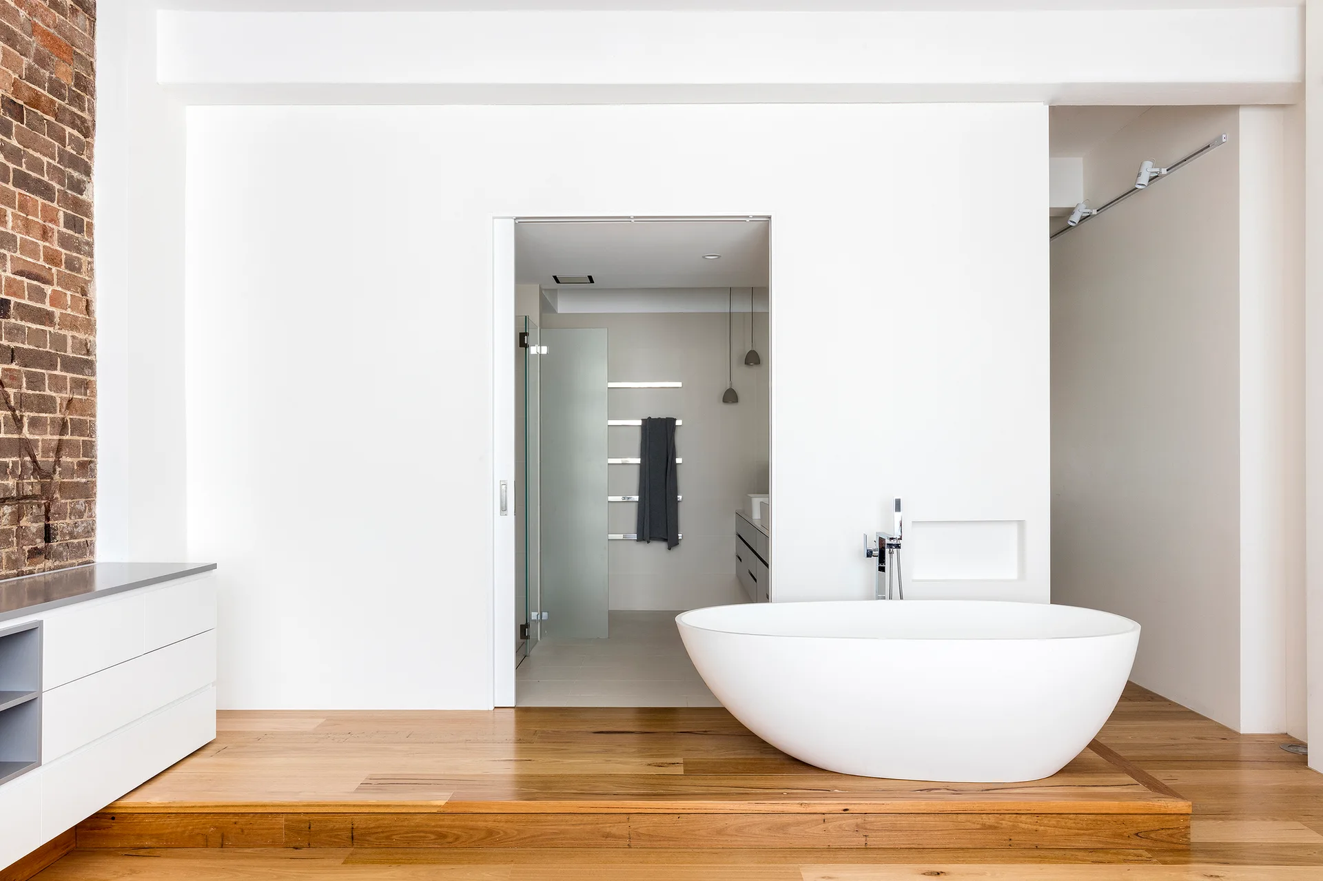A white freestanding bathtub sits on a raised wooden platform in a minimalist bathroom with white walls and a glimpse of an exposed brick wall.