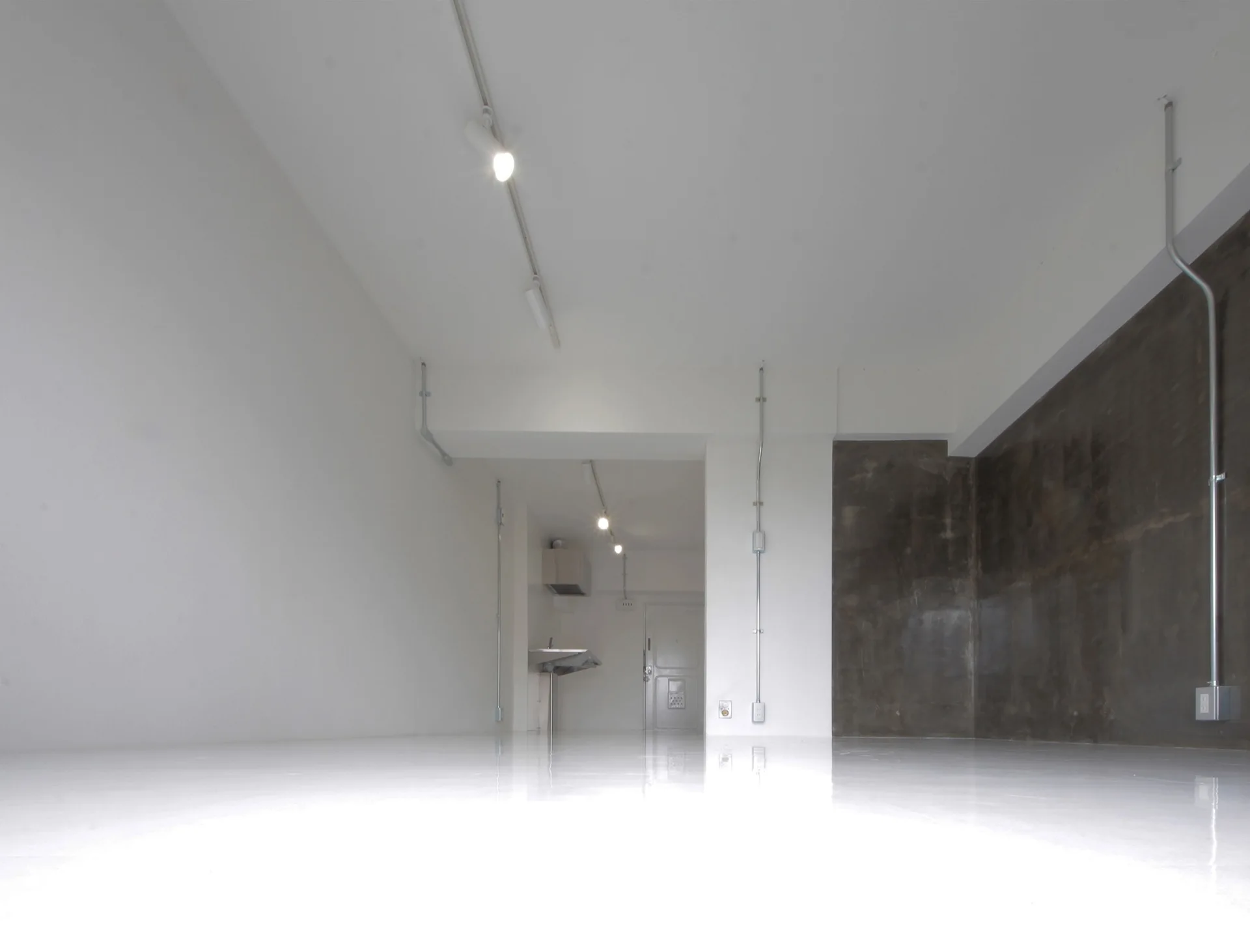 An empty industrial minimalist room featuring high-gloss white floors, a dark concrete accent wall, and exposed metal conduit pipes.