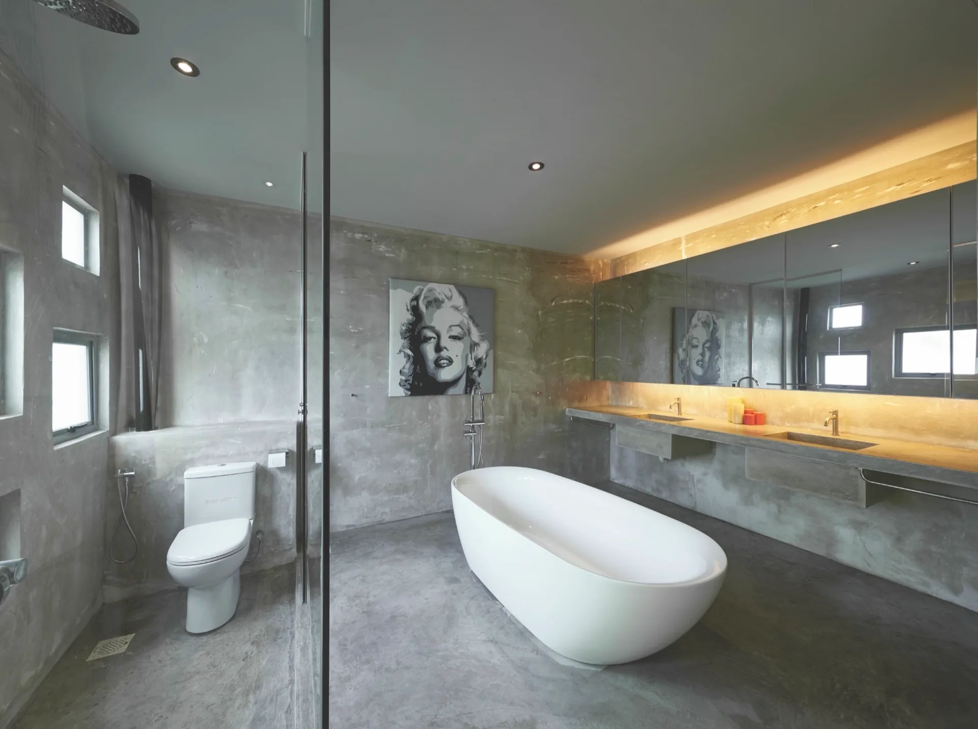 A minimalist bathroom with raw concrete walls and floors, featuring a white freestanding bathtub and a long wall-mounted vanity under a large mirror.