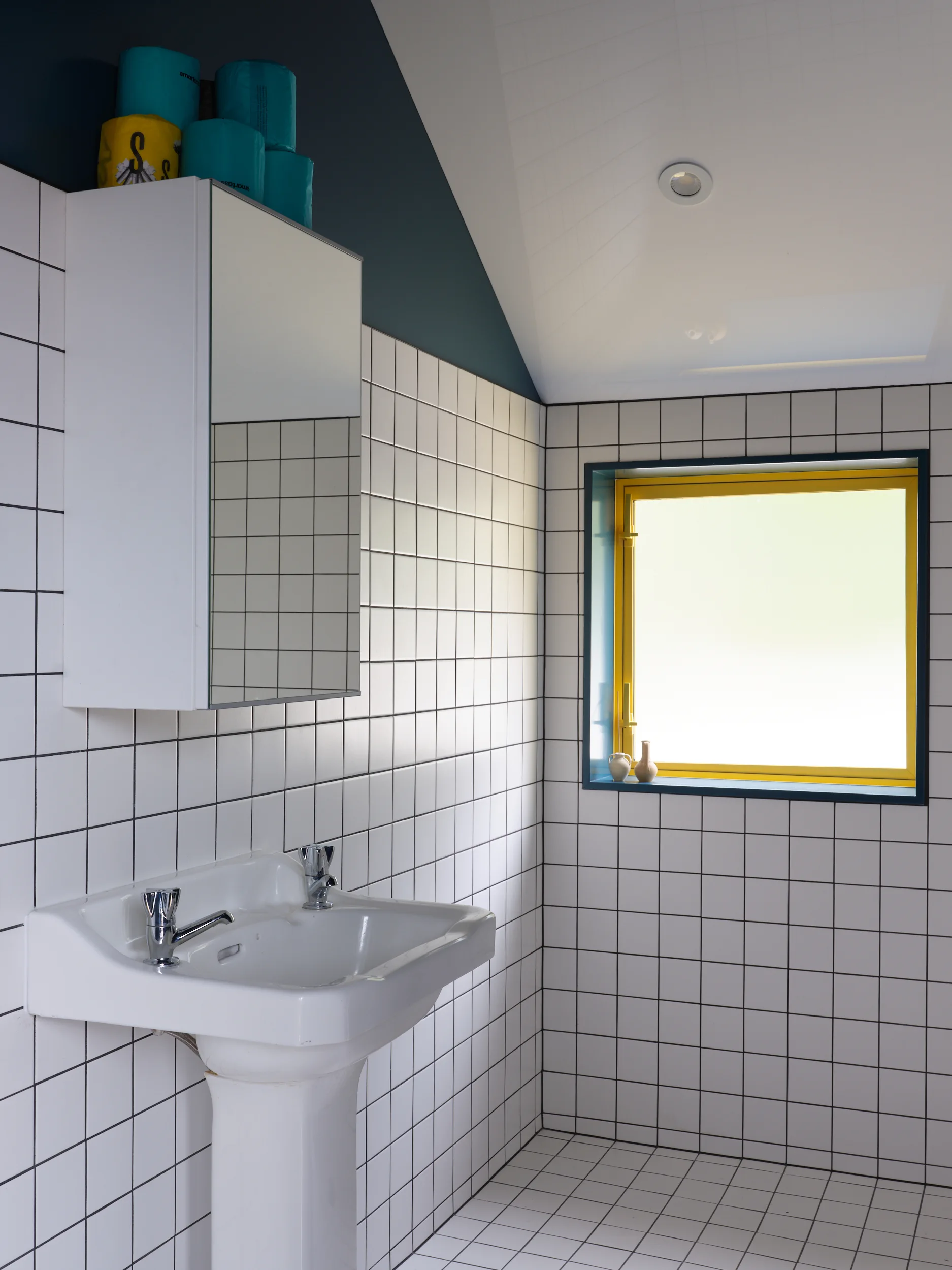 A modern bathroom featuring white square grid tiled walls and floor, a white pedestal sink, and a high-contrast window with a bright yellow frame.