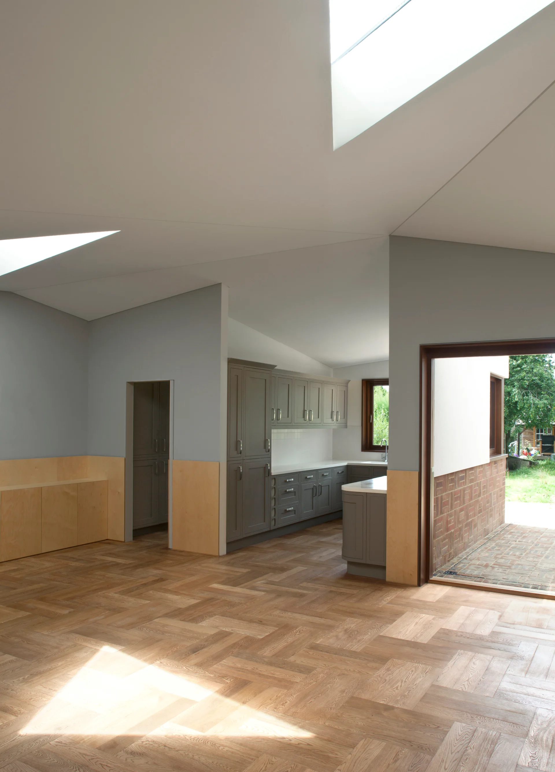 A minimalist open-plan kitchen featuring grey cabinetry, natural light wood herringbone flooring, wall paneling, and a large geometric skylight.
