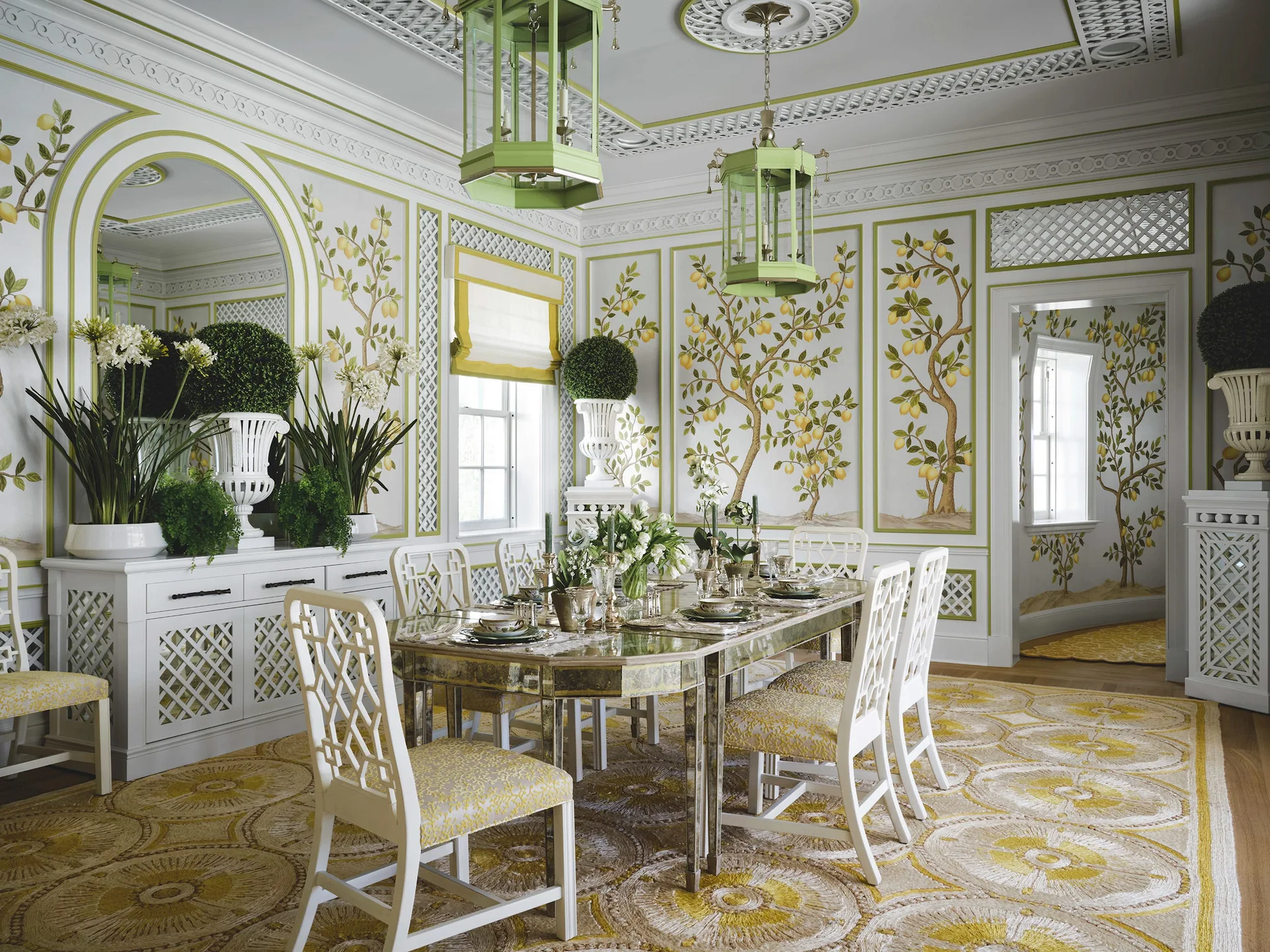 A formal dining room features a mirrored dining table surrounded by white lattice chairs, under hanging green lantern lights and framed lemon wallpaper.