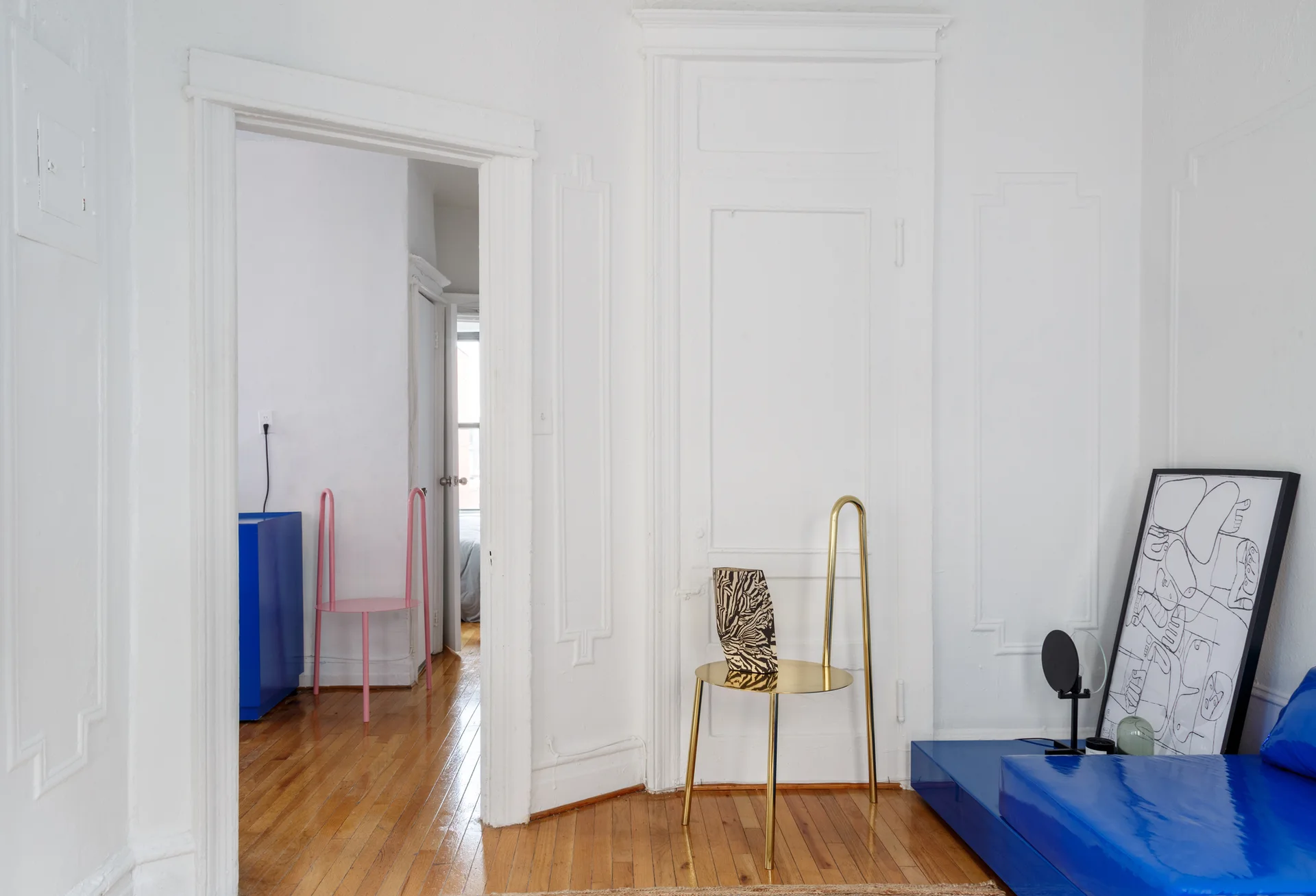 A bright room with white paneled walls and hardwood floors features a gold metal chair, a low blue sofa, and a framed sketch leaning on the floor.