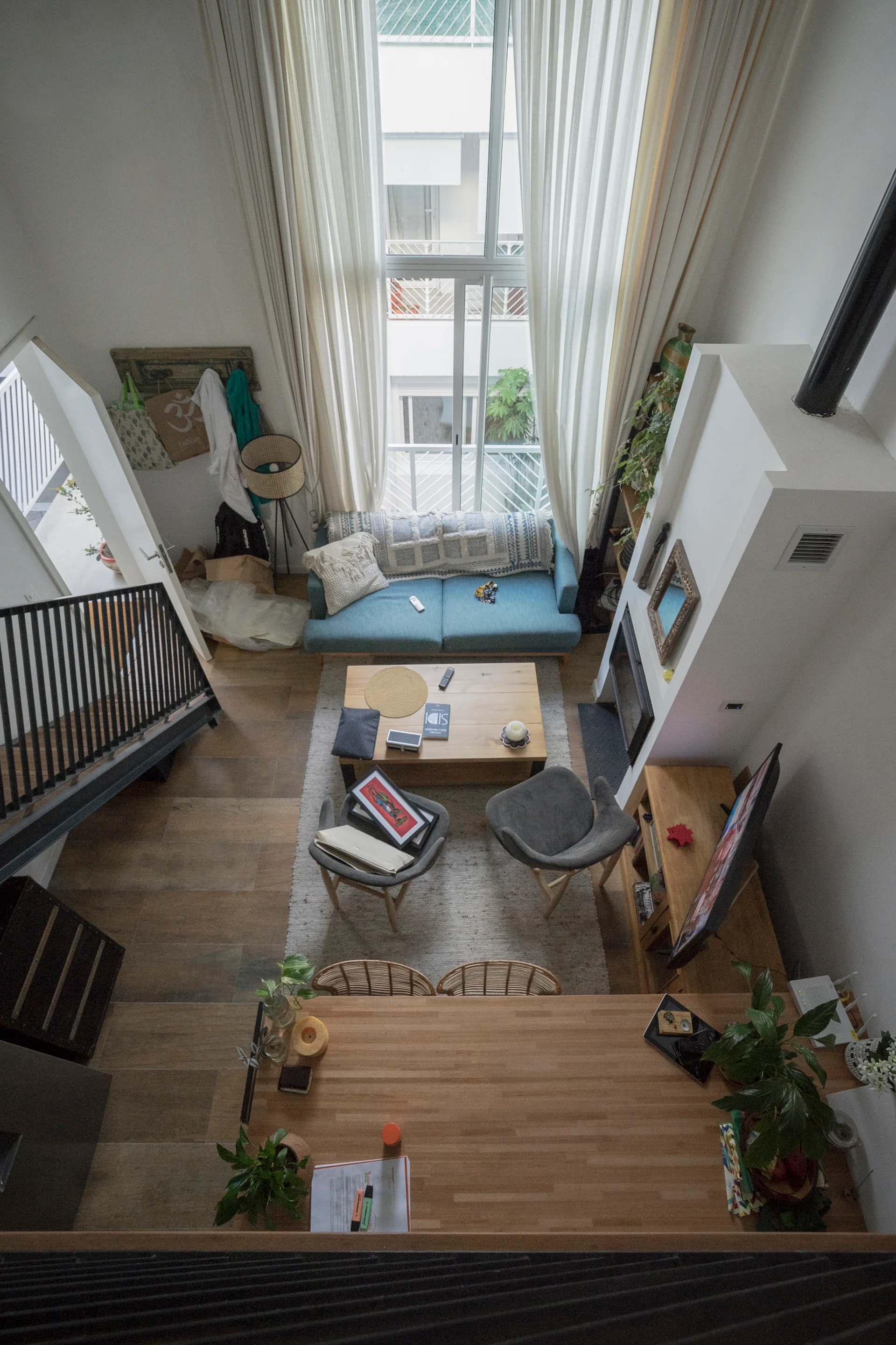 A high-angle view of a double-height living room featuring a blue sofa, wooden coffee table, two gray armchairs, and tall windows with sheer curtains.