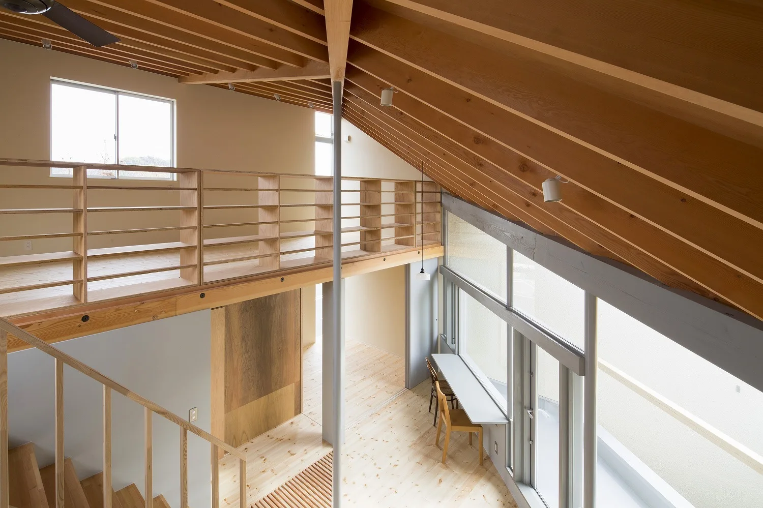A minimalist modern loft interior featuring a wooden mezzanine landing, an angled rafter ceiling, and a desk built along a wall of large windows.