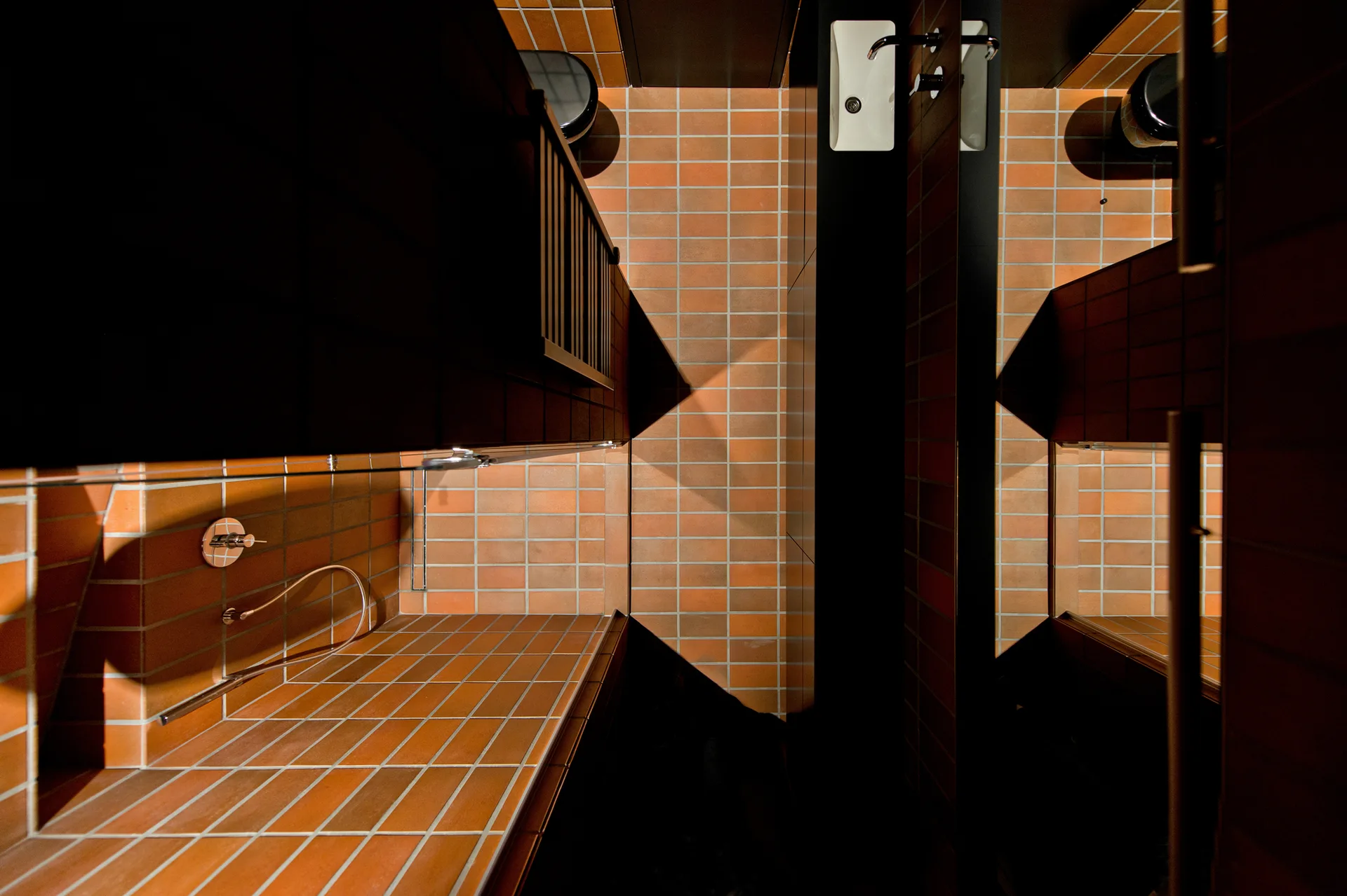 A minimalist bathroom viewed from above, featuring terracotta square tiles, a white integrated sink on a black vanity, and a chrome faucet.