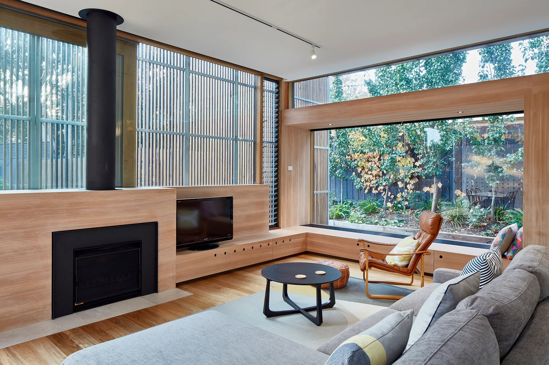 A modern living room features a grey sectional sofa, a round coffee table, a wooden fireplace surround, and a low bench beneath large windows.