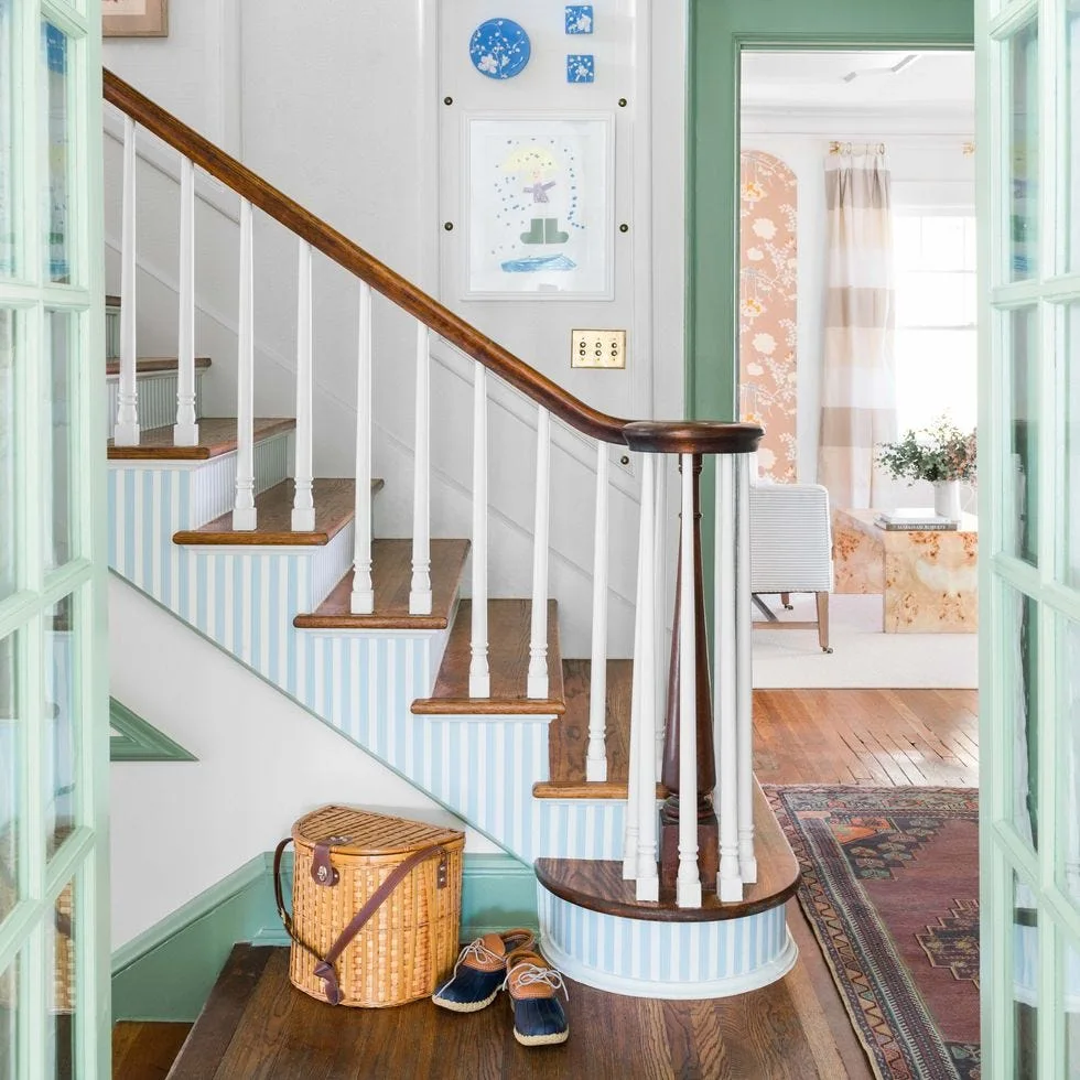 A wooden staircase features blue and white striped risers, white spindles, and a dark wood handrail next to an open doorway framed in sage green trim.