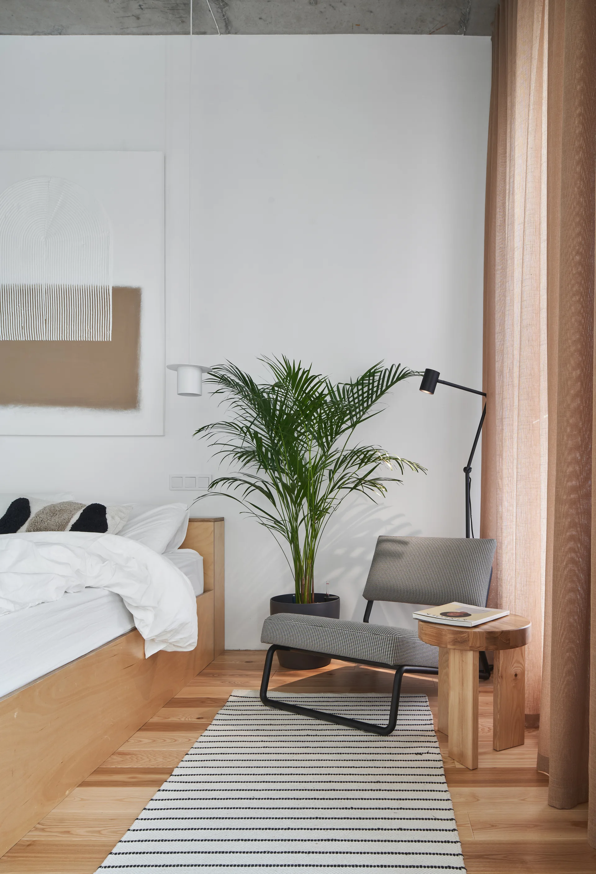 A minimalist bedroom featuring a light wood platform bed, a houndstooth armchair, and a green potted palm against a white wall.