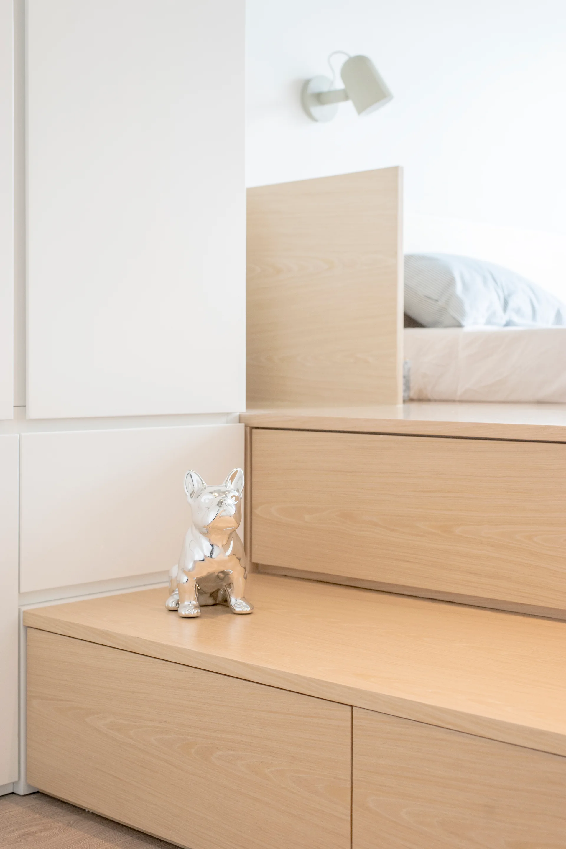 A minimalist bedroom features light wood platform bed steps integrated with white built-in cabinetry and a small metallic bulldog figurine.