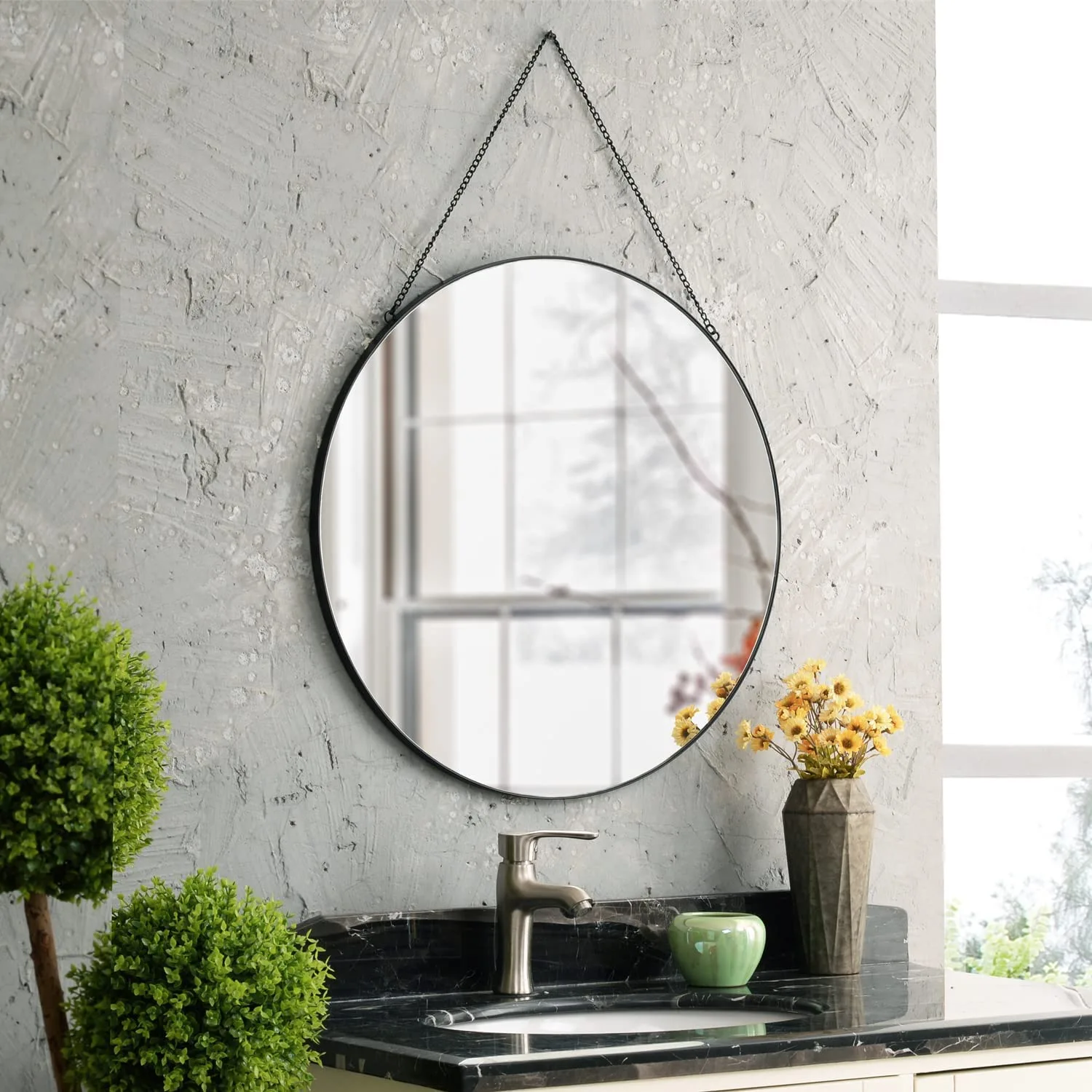 A circular black-framed mirror hangs by a chain against a grey plaster wall above a black marble vanity with a metal faucet.