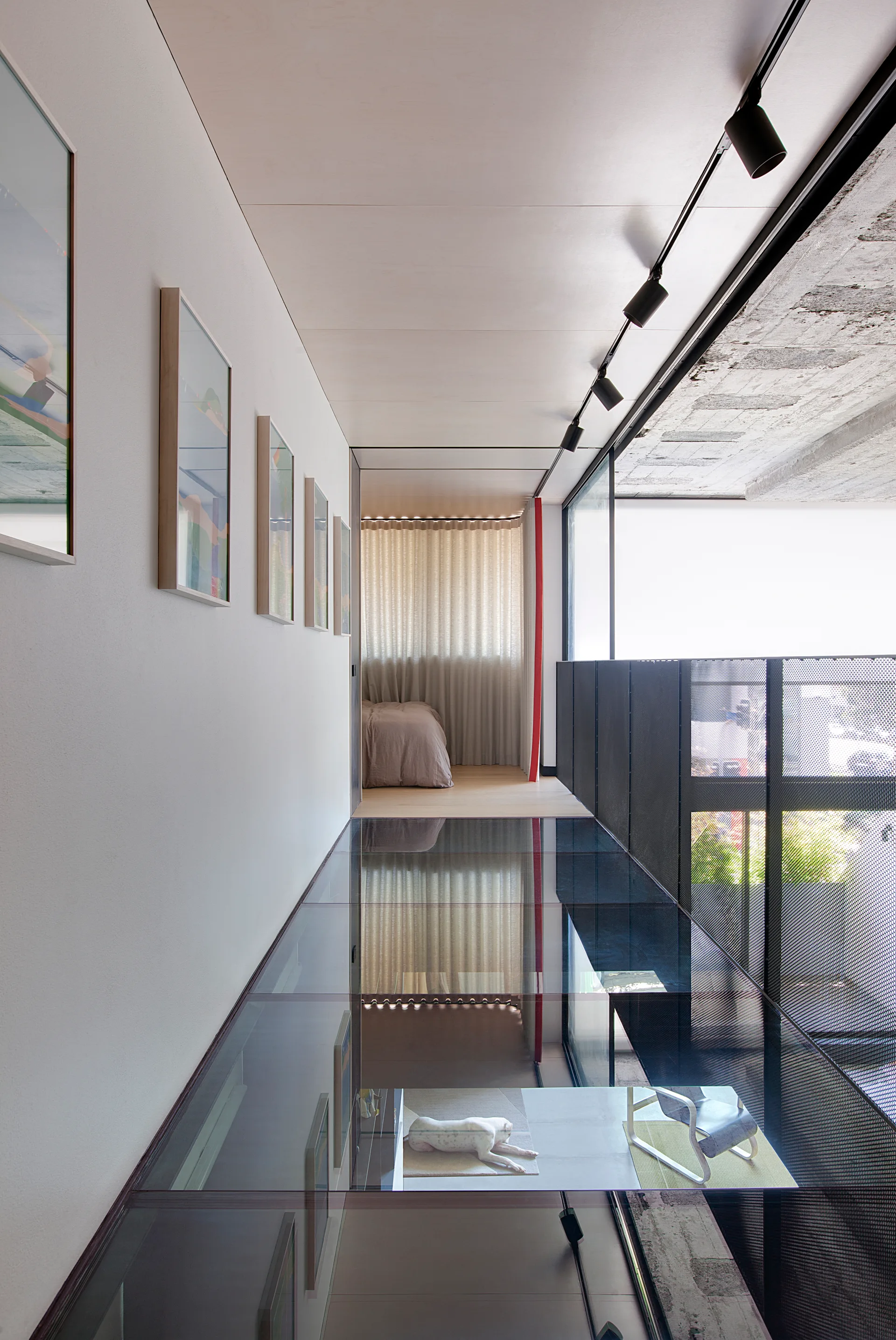 A modern hallway features a transparent glass floor overlooking a lower level, a white wall with framed artwork, and black track lighting.