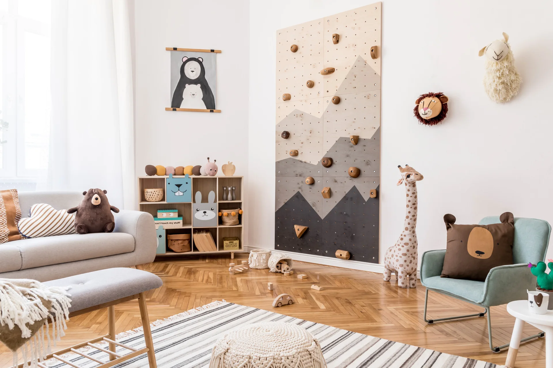 A bright kids' playroom featuring a mountain-themed climbing wall, a wooden cubby shelf, a gray sofa, and light herringbone wood floors.
