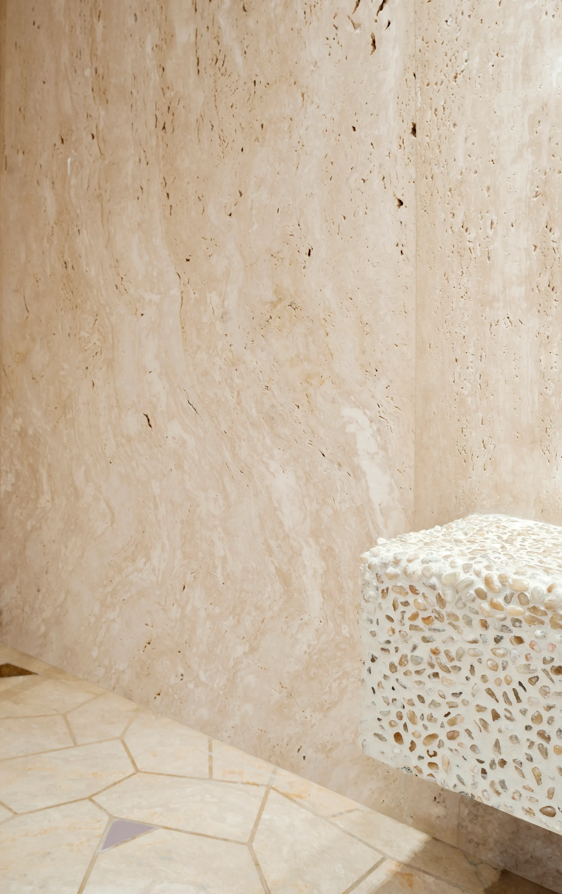 A detailed close-up of a beige travertine stone wall, a geometric tiled floor, and a rectangular block textured with small embedded pebbles.