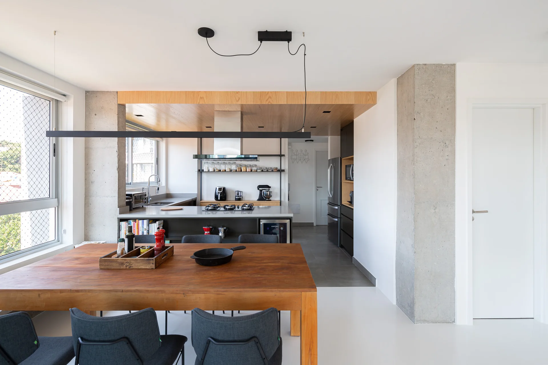 An open-concept kitchen and dining room featuring a wooden table under a black linear pendant light, exposed concrete pillars, and a wood-paneled ceiling.