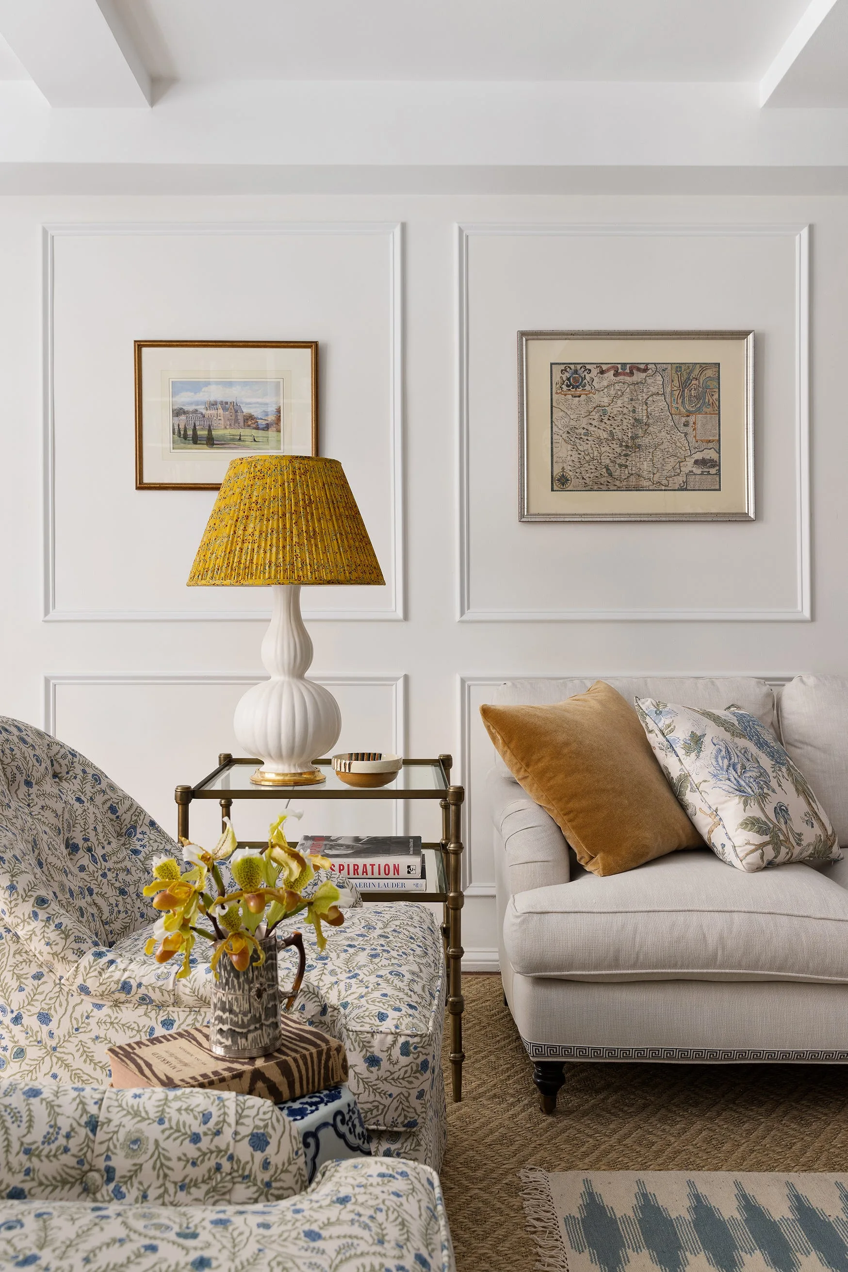 A traditional living room corner featuring a floral armchair, a cream sofa with decorative pillows, and a yellow table lamp against paneled walls.