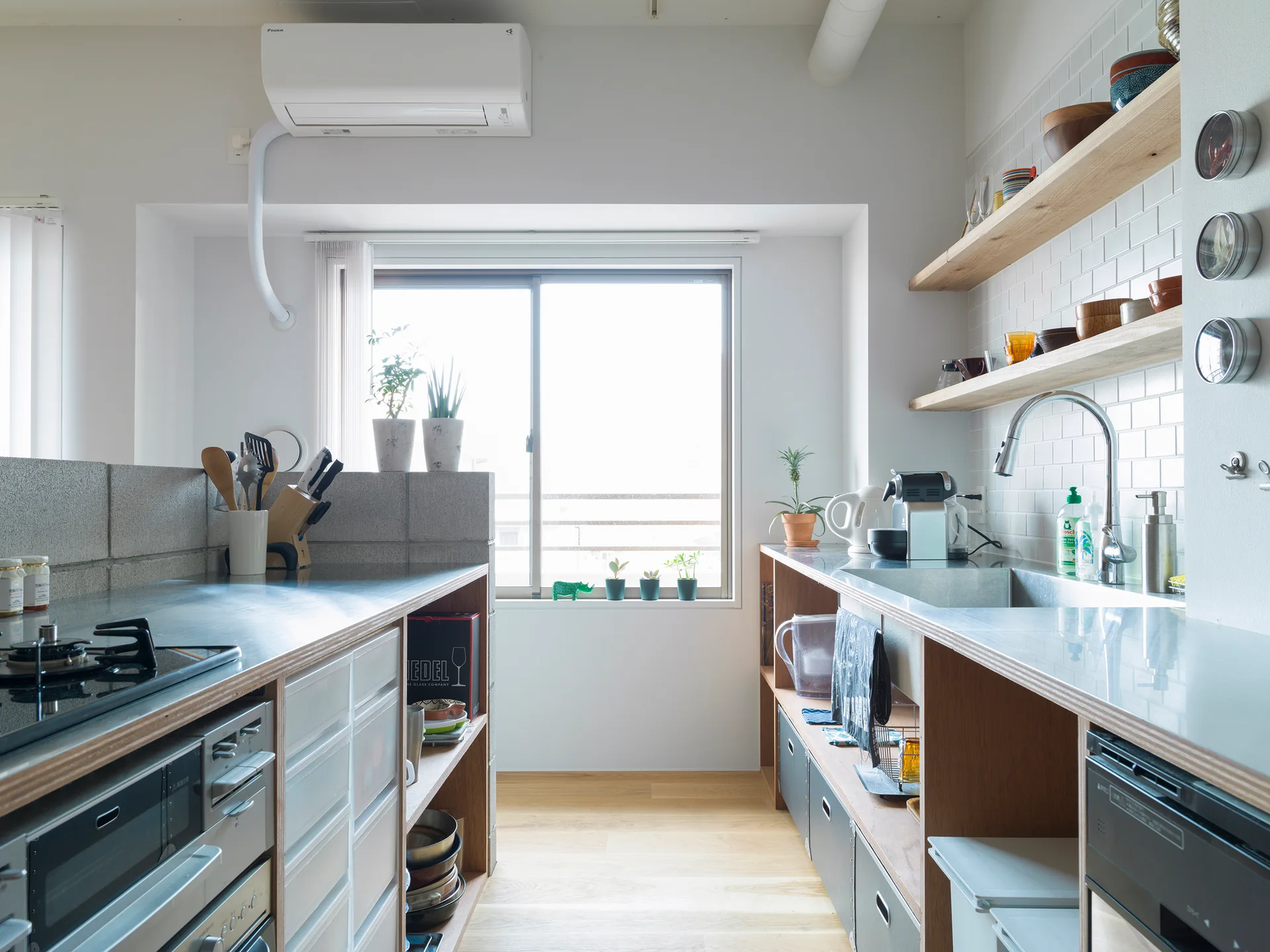 A galley kitchen features stainless steel countertops, open wooden shelving on a white tiled wall, and a concrete block partition beside a bright window.