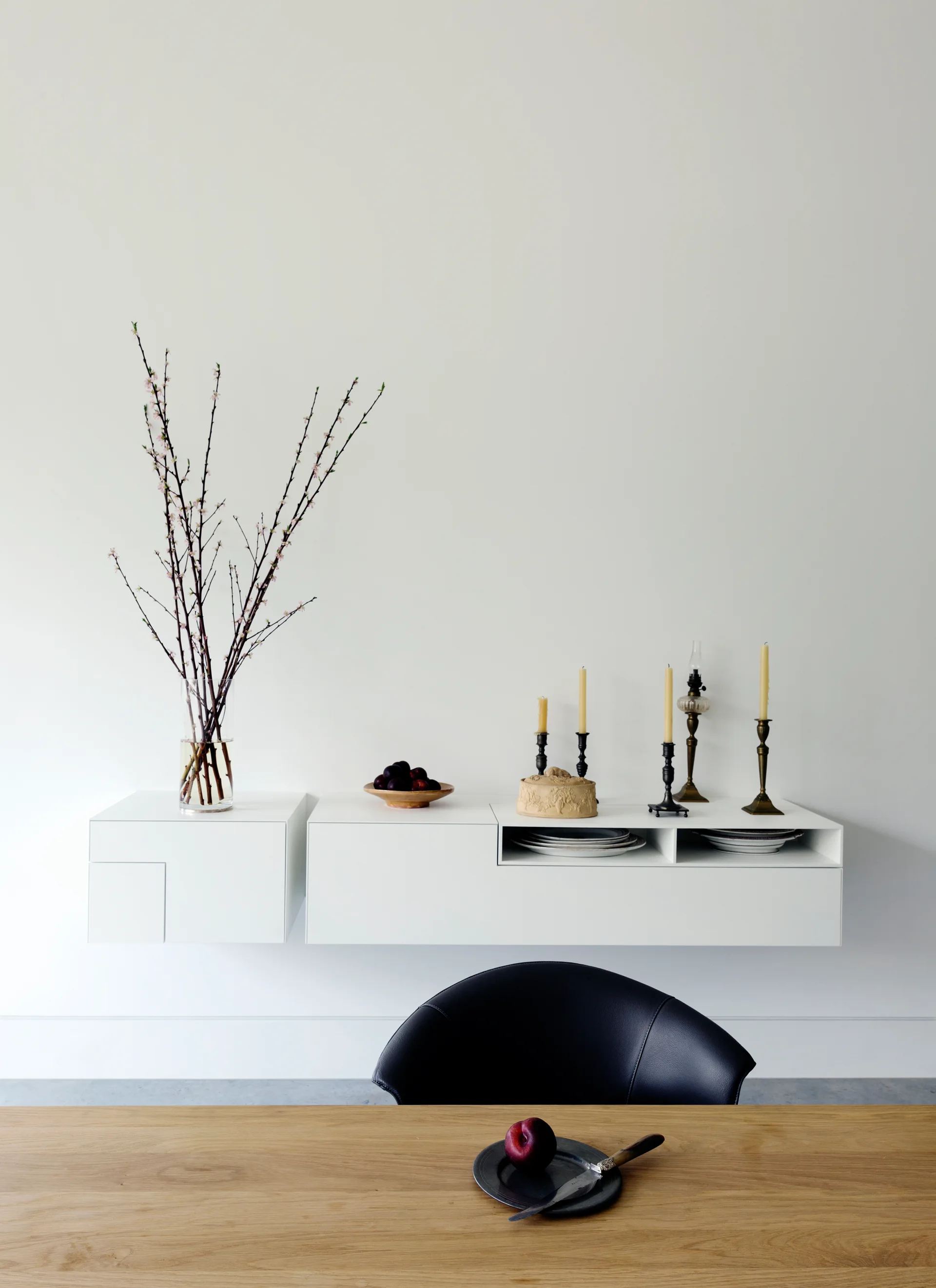 A minimalist white wall features a floating white cabinet adorned with tall branches and candlesticks above a wood table.