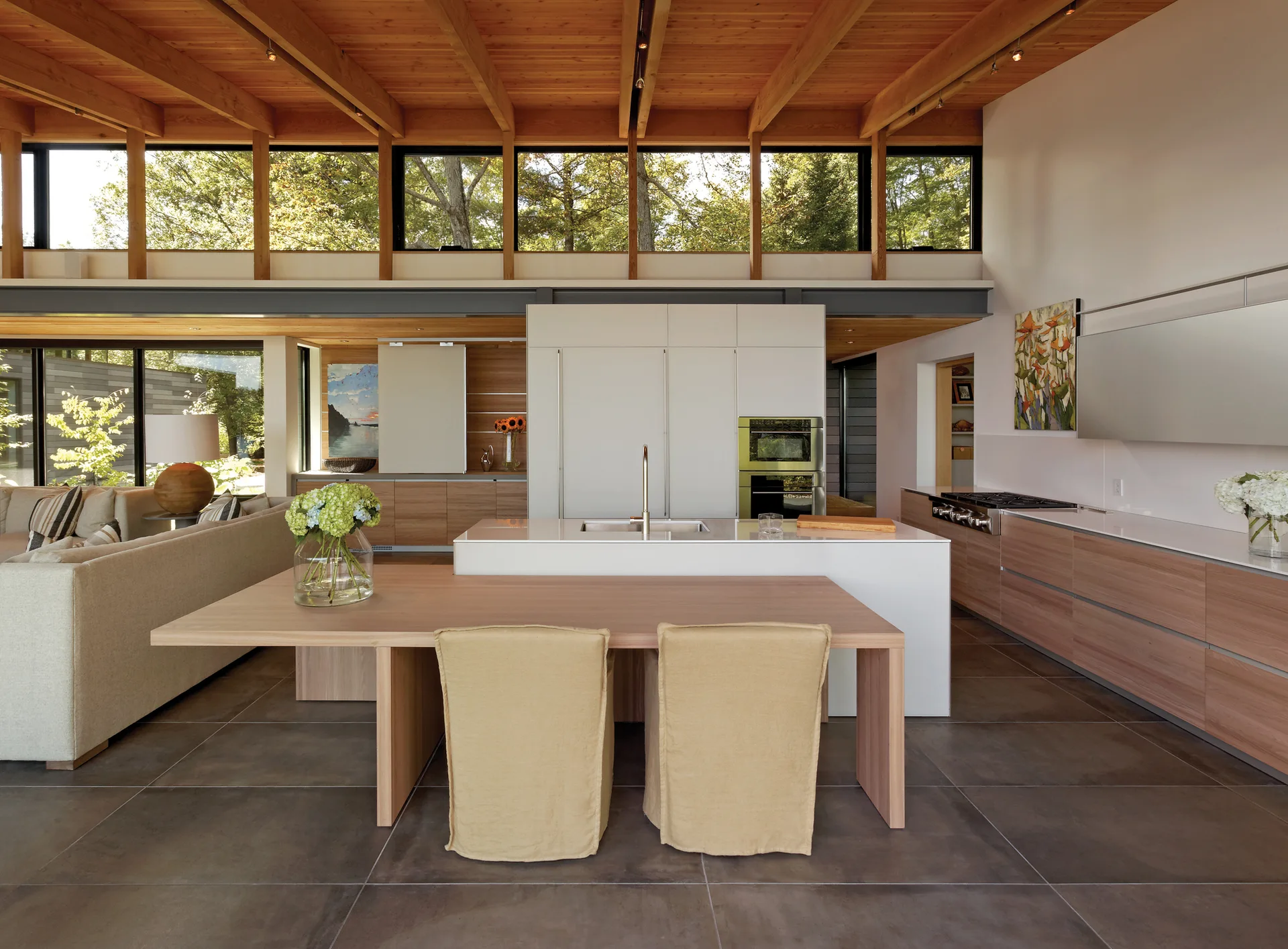 An open kitchen features a central white island, integrated wood dining table with slipcovered chairs, wood-clad ceiling, and high clerestory windows.