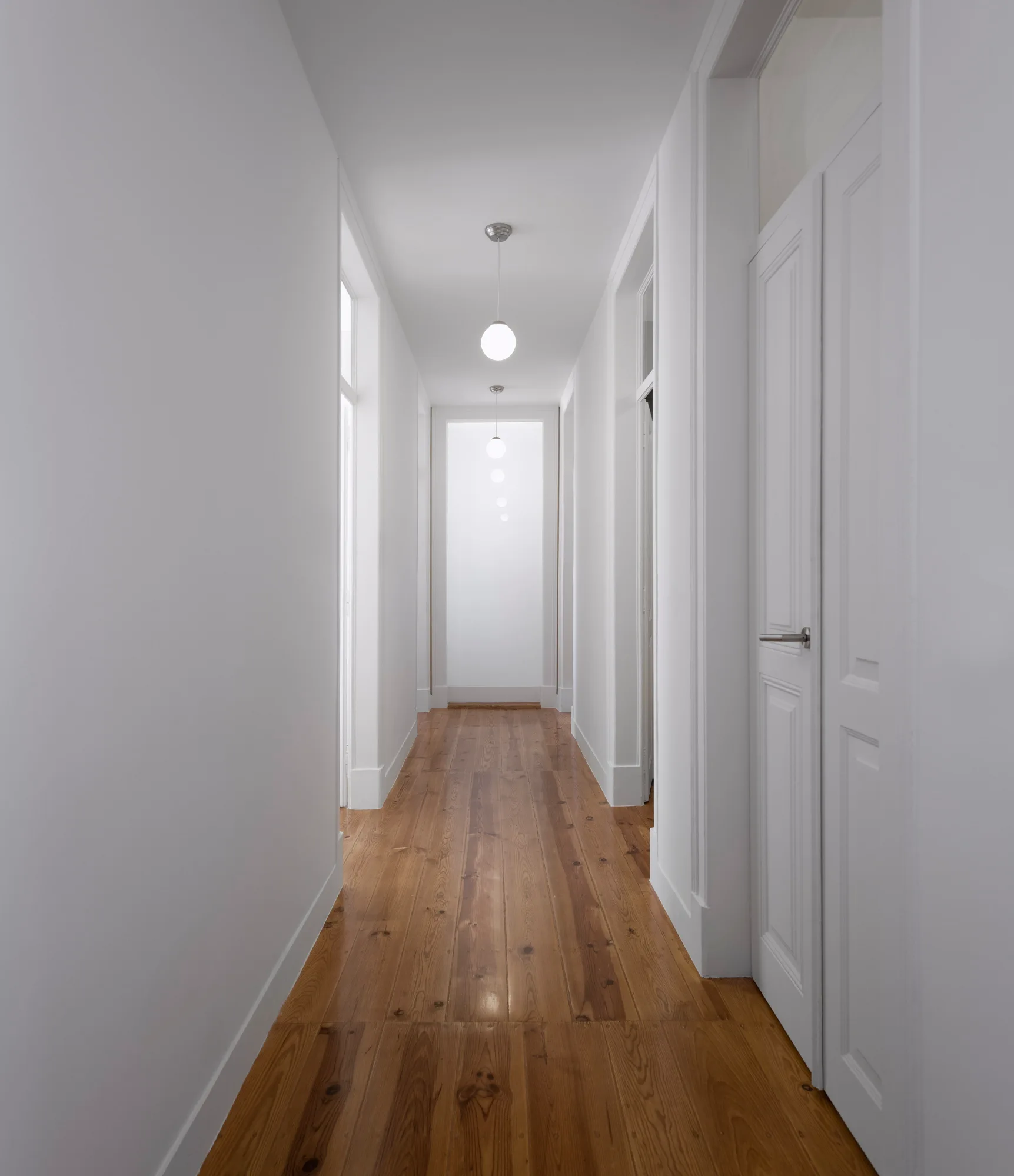 A long, narrow white hallway features natural wood plank flooring, white paneled doors, and a line of hanging globe pendant lights.