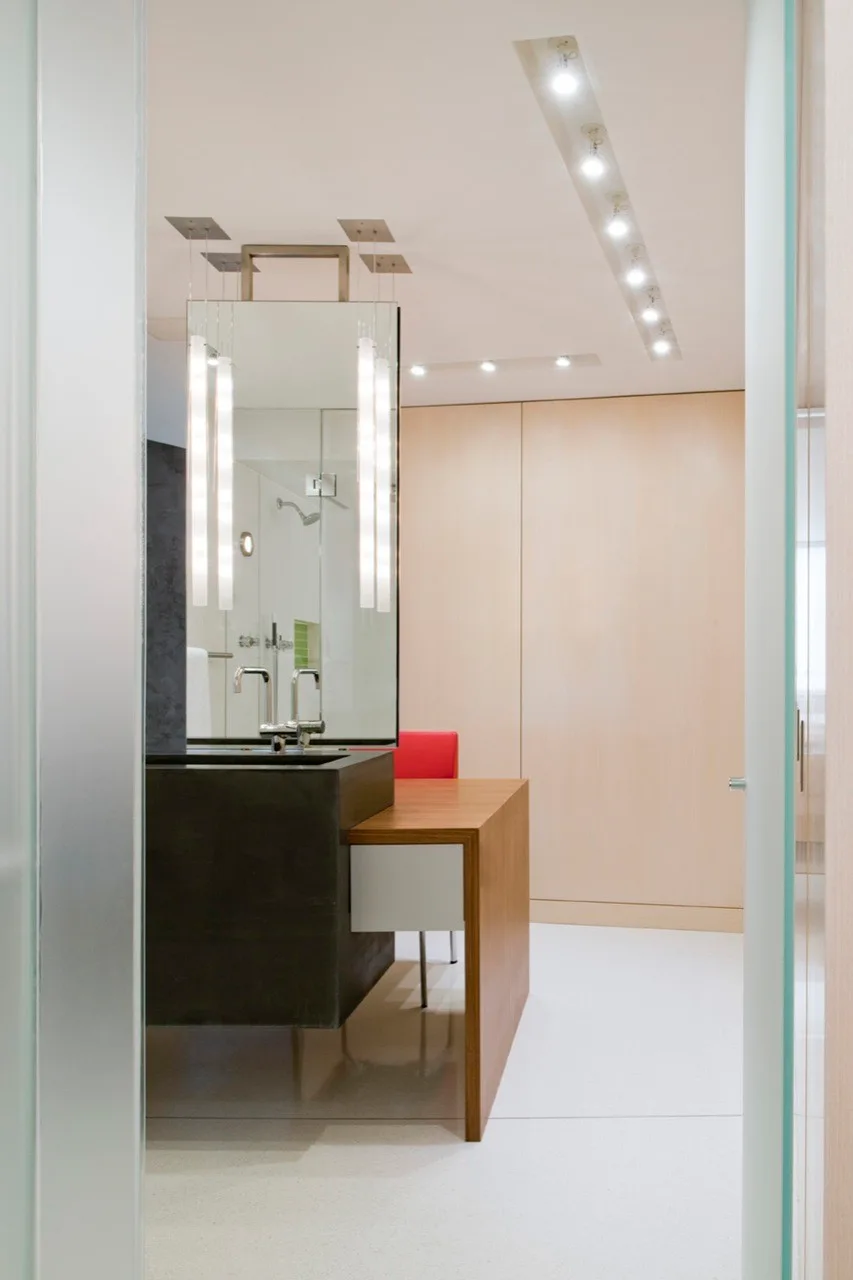 A modern bathroom features a dark concrete vanity with an attached wooden desk, a tall vertical mirror with pendant lights, and light wood wall paneling.