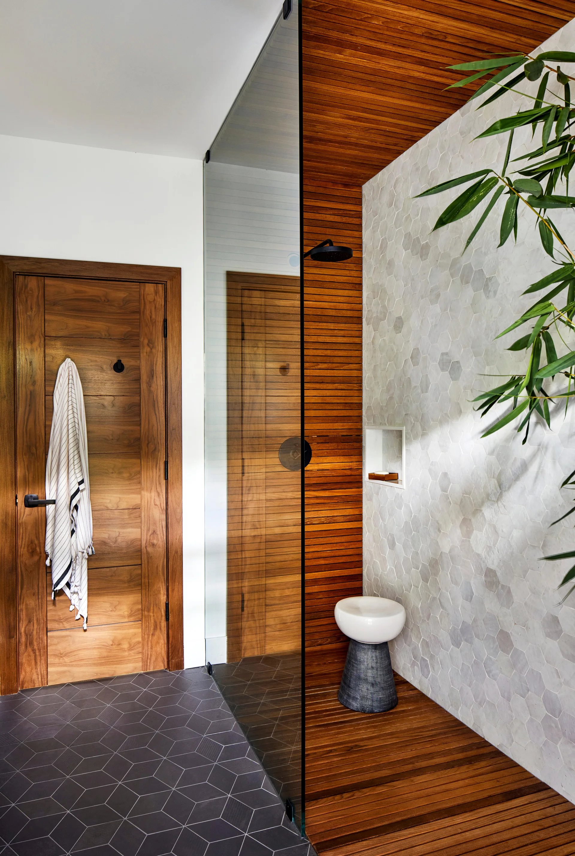 A modern bathroom features a glass-enclosed shower with horizontal wood slats, light grey hexagonal wall tiles, and a natural wood door.
