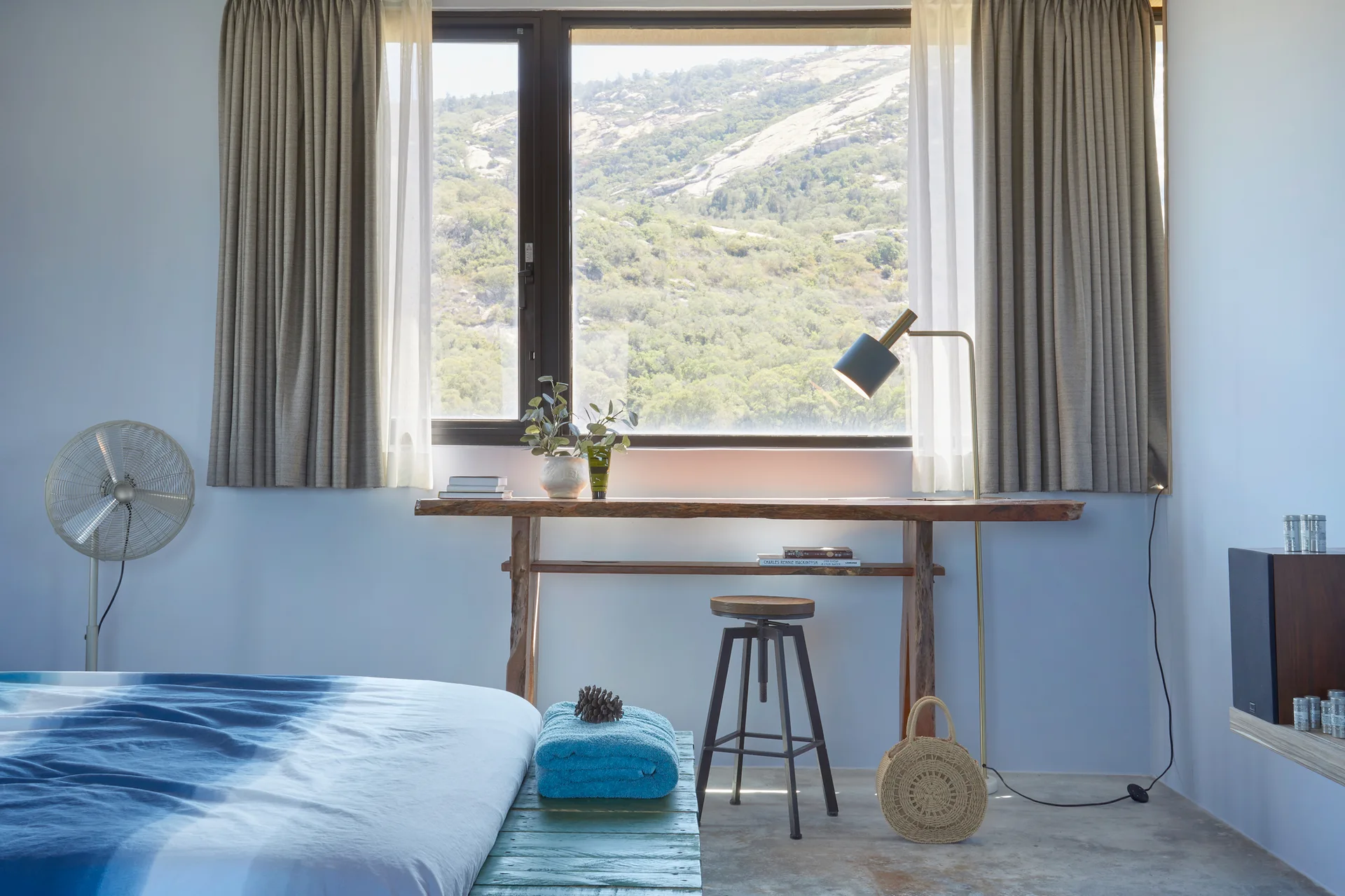 A minimalist bedroom featuring a live-edge wooden desk placed under a large window with mountain views, framed by neutral drapes.