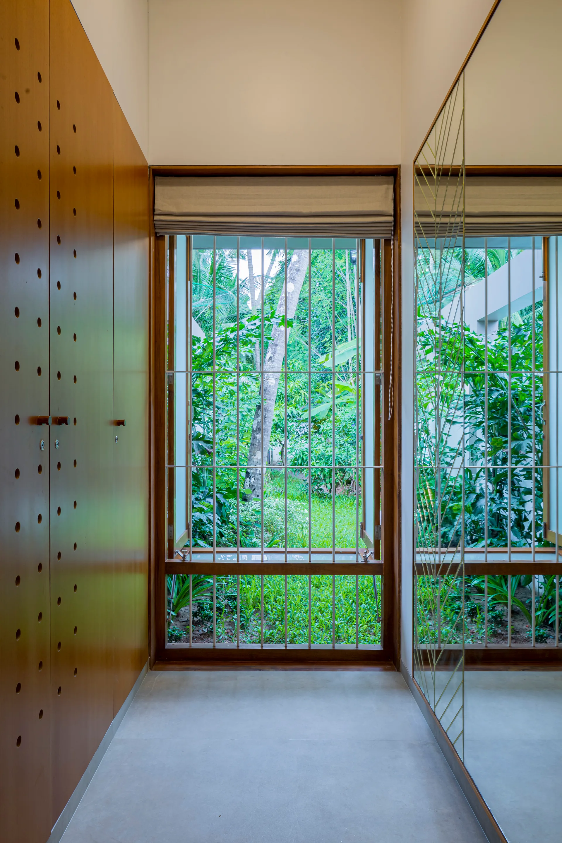 A narrow hallway features a perforated wood wardrobe on one side and a large etched mirror reflecting a window with a lush green garden view.