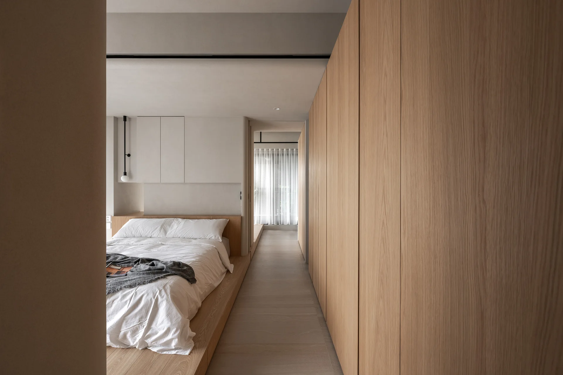 A minimalist bedroom featuring a low wood platform bed and a wall of built-in light oak cabinets leading toward a window with sheer curtains.