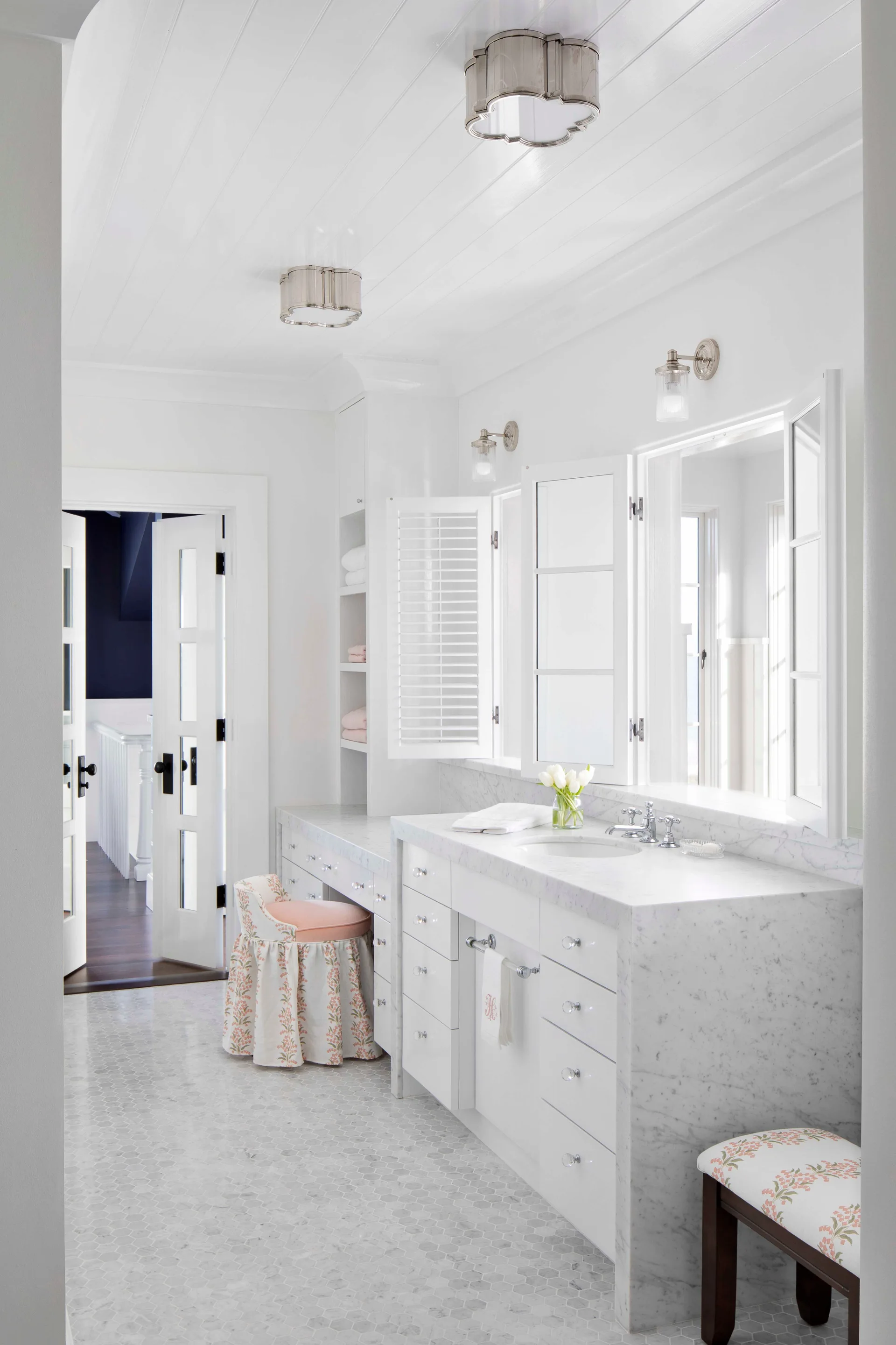 A bright white bathroom features a marble-topped vanity with crystal knobs, a skirted floral stool, and small hexagonal marble floor tiles.