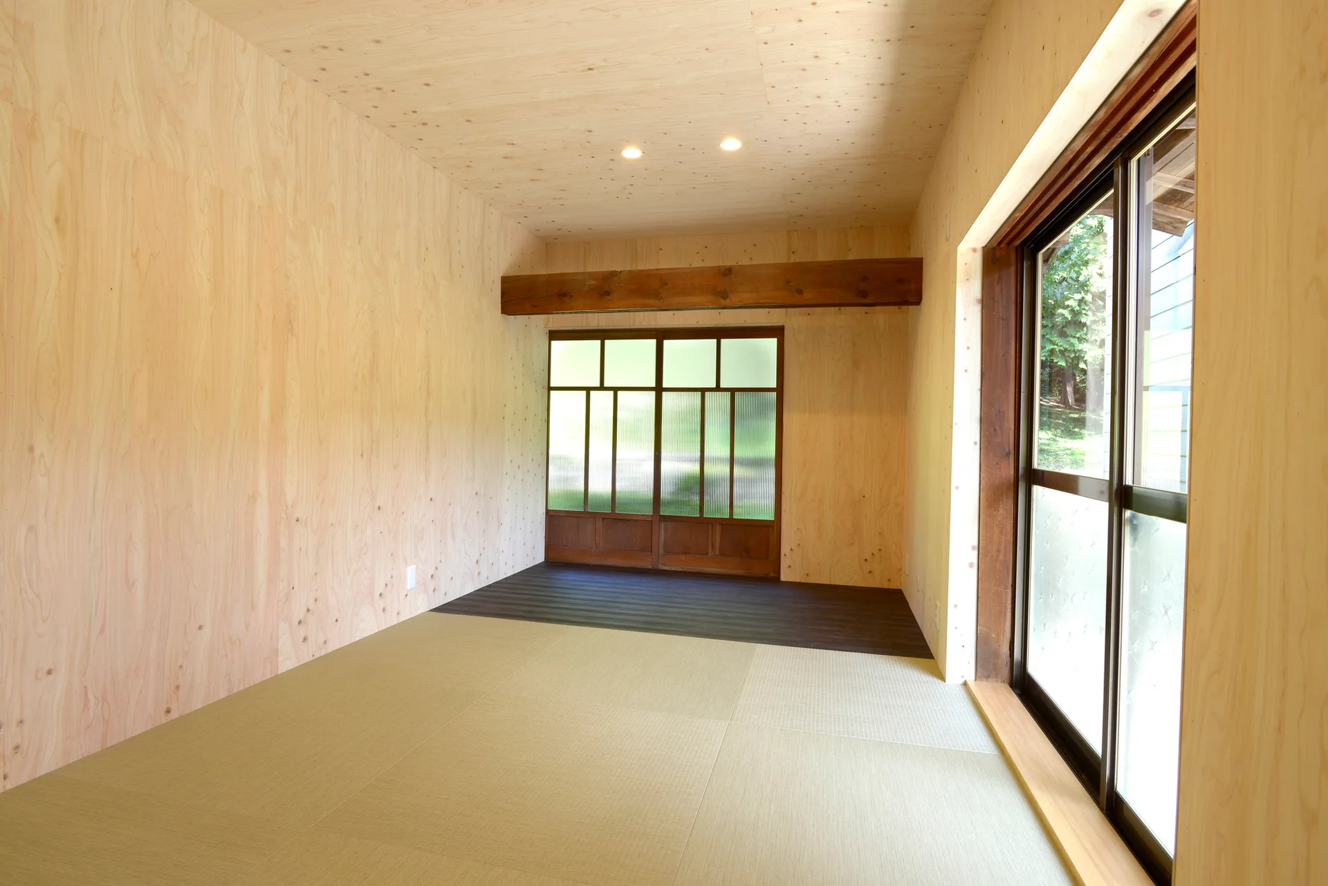 An empty Japanese modern room features light plywood walls and ceiling with tatami mat flooring, a timber beam, and sliding doors.