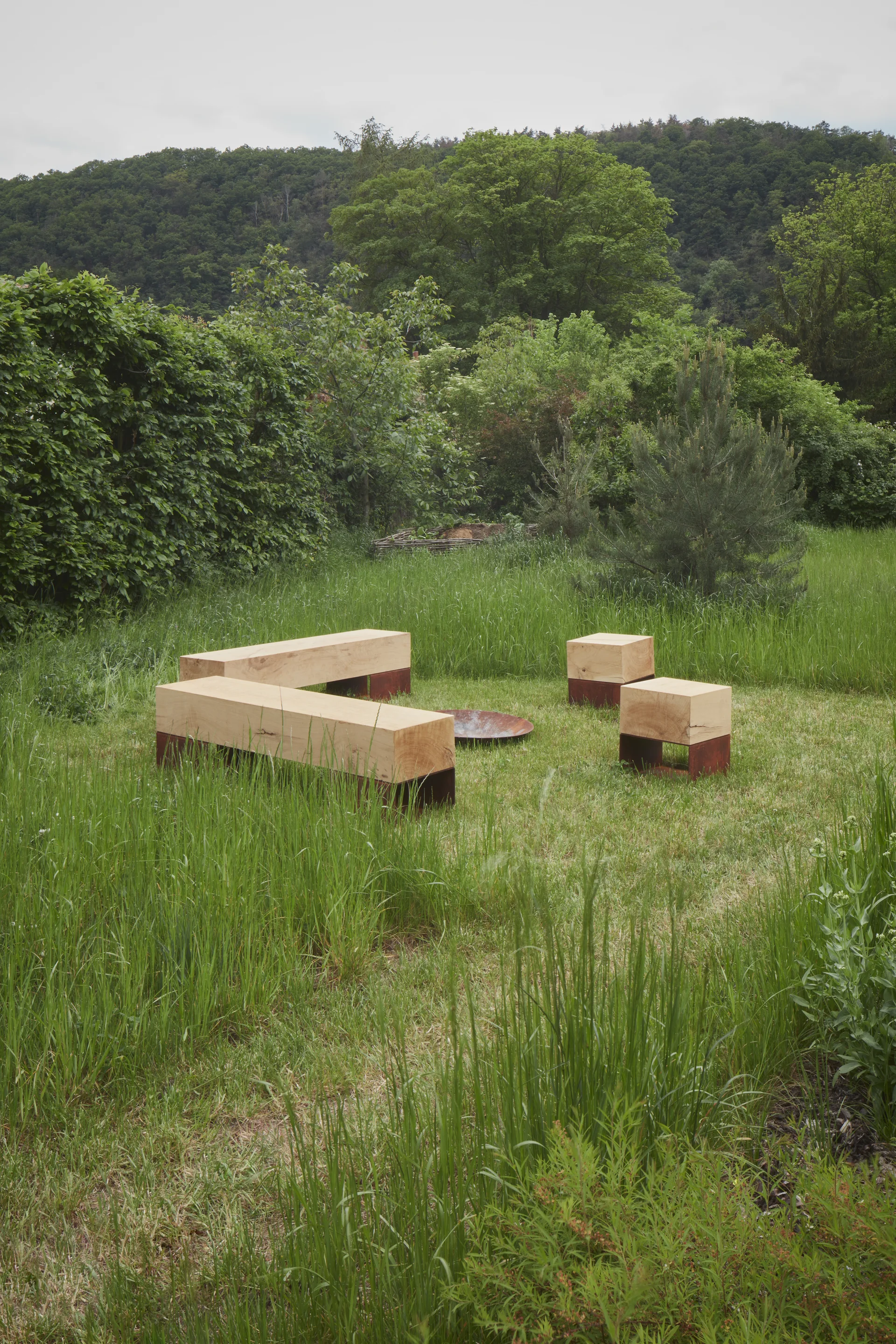 Minimalist wooden block benches and cube stools are arranged around a circular metal fire pit in a grassy outdoor meadow.