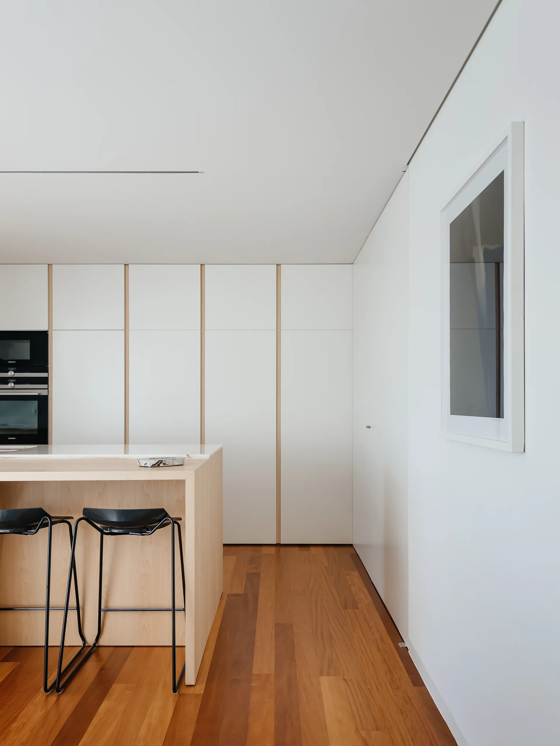 A minimalist kitchen features a light wood island with black stools, seamless white floor-to-ceiling cabinets, and hardwood floors.
