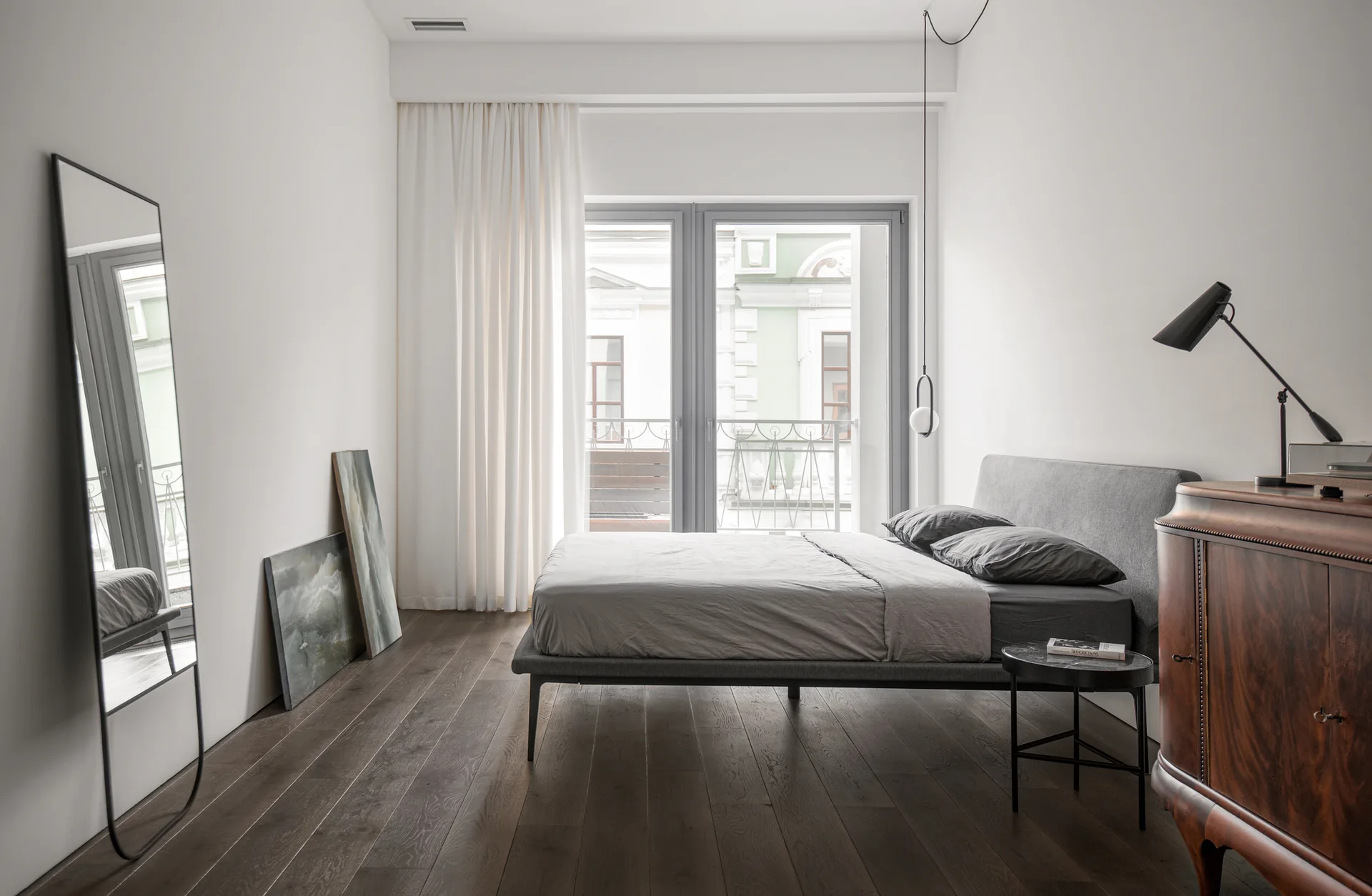 A minimalist bedroom featuring a grey bed on dark wood floors, a vintage wooden cabinet, and a tall floor mirror against white walls.