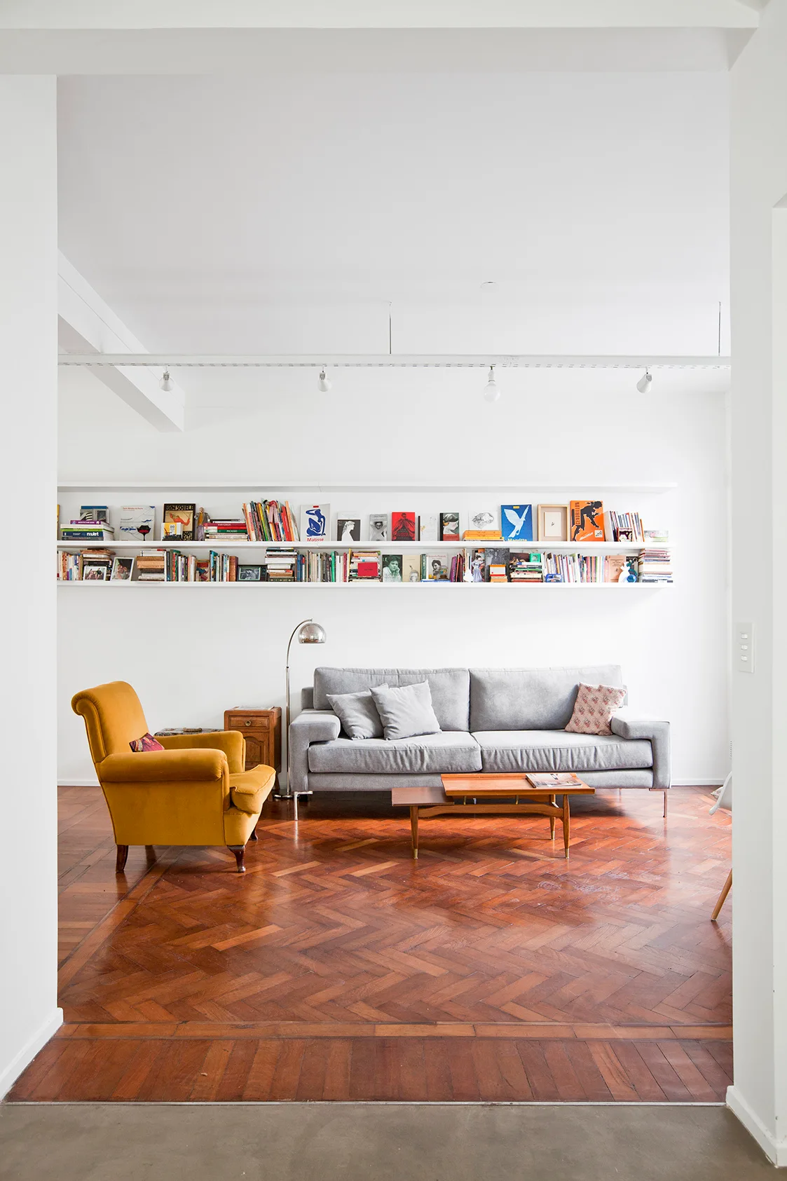 A modern living room features a grey sofa, a mustard yellow armchair, and long white floating bookshelves over a warm herringbone parquet wood floor.