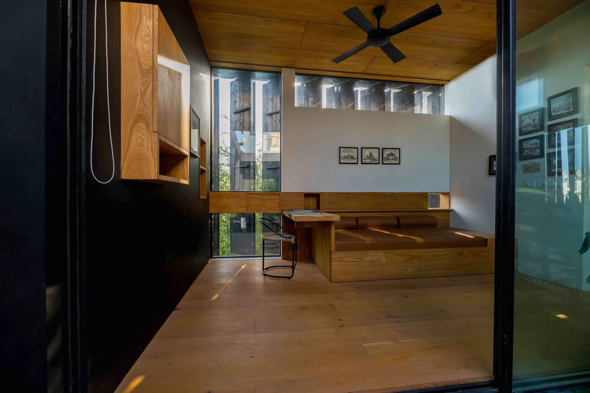 A minimalist bedroom featuring a custom built-in wooden daybed and desk unit, a wood plank ceiling with a black ceiling fan, and a matte black wall.